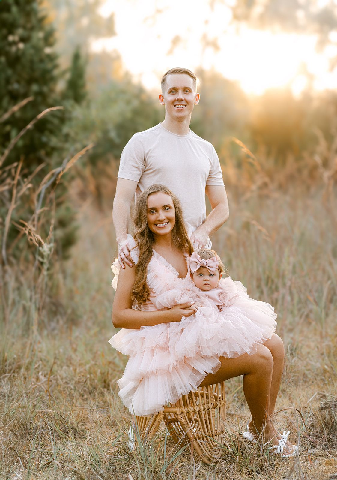 Family portrait of couple with baby in pink tutu sitting in wicker chair in sunlit autumn meadow.