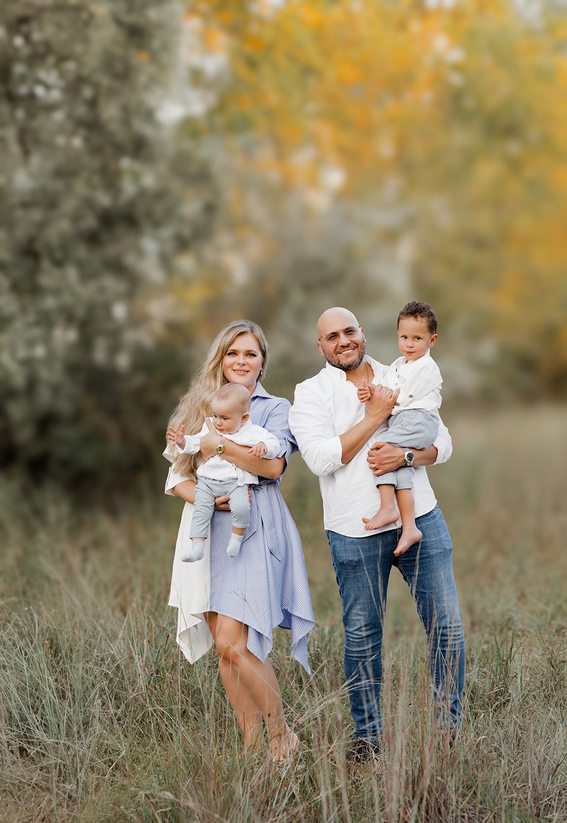 A smiling family of four poses in an autumn field, parents each holding a young child in casual attire.