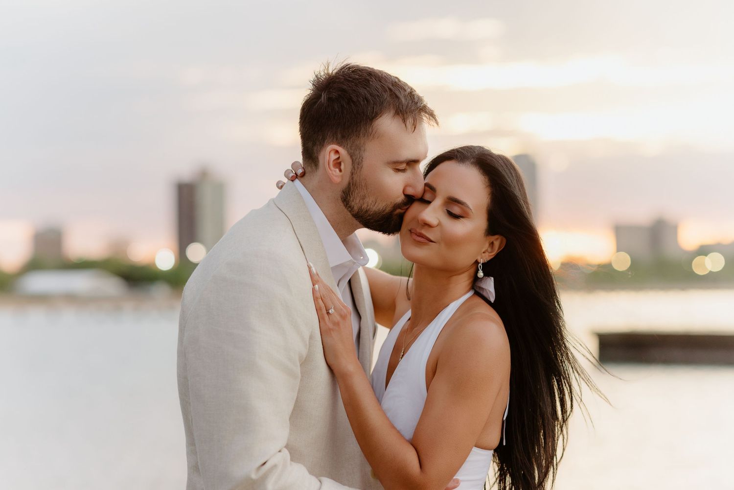 Man kissing woman on cheek during sunset engagement photo session by city waterfront.