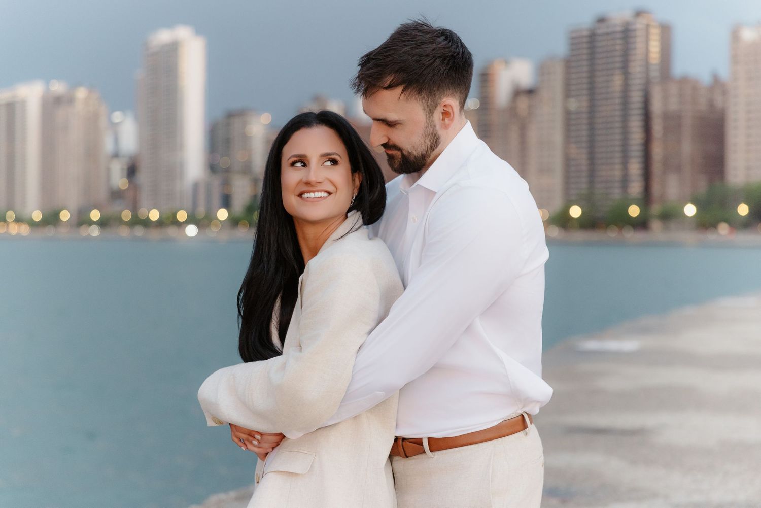 Couple embracing on Chicago waterfront with city skyline bokeh backdrop during golden hour engagement photos.
