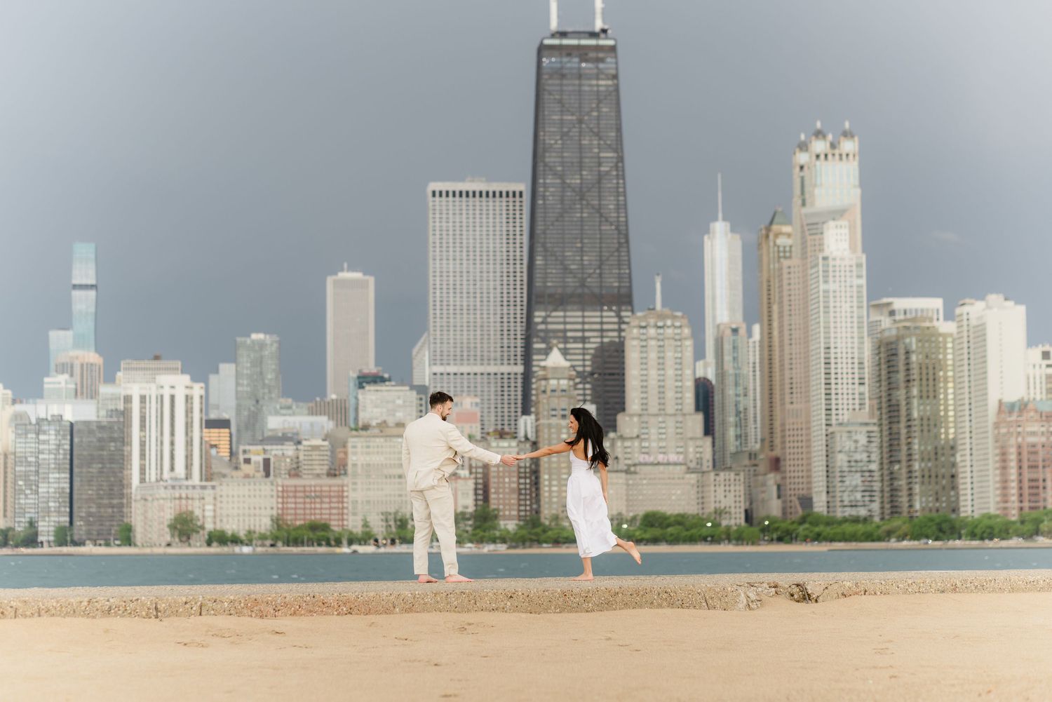 Newly engaged couple holding hands on Chicago beach with iconic John Hancock Tower skyline.