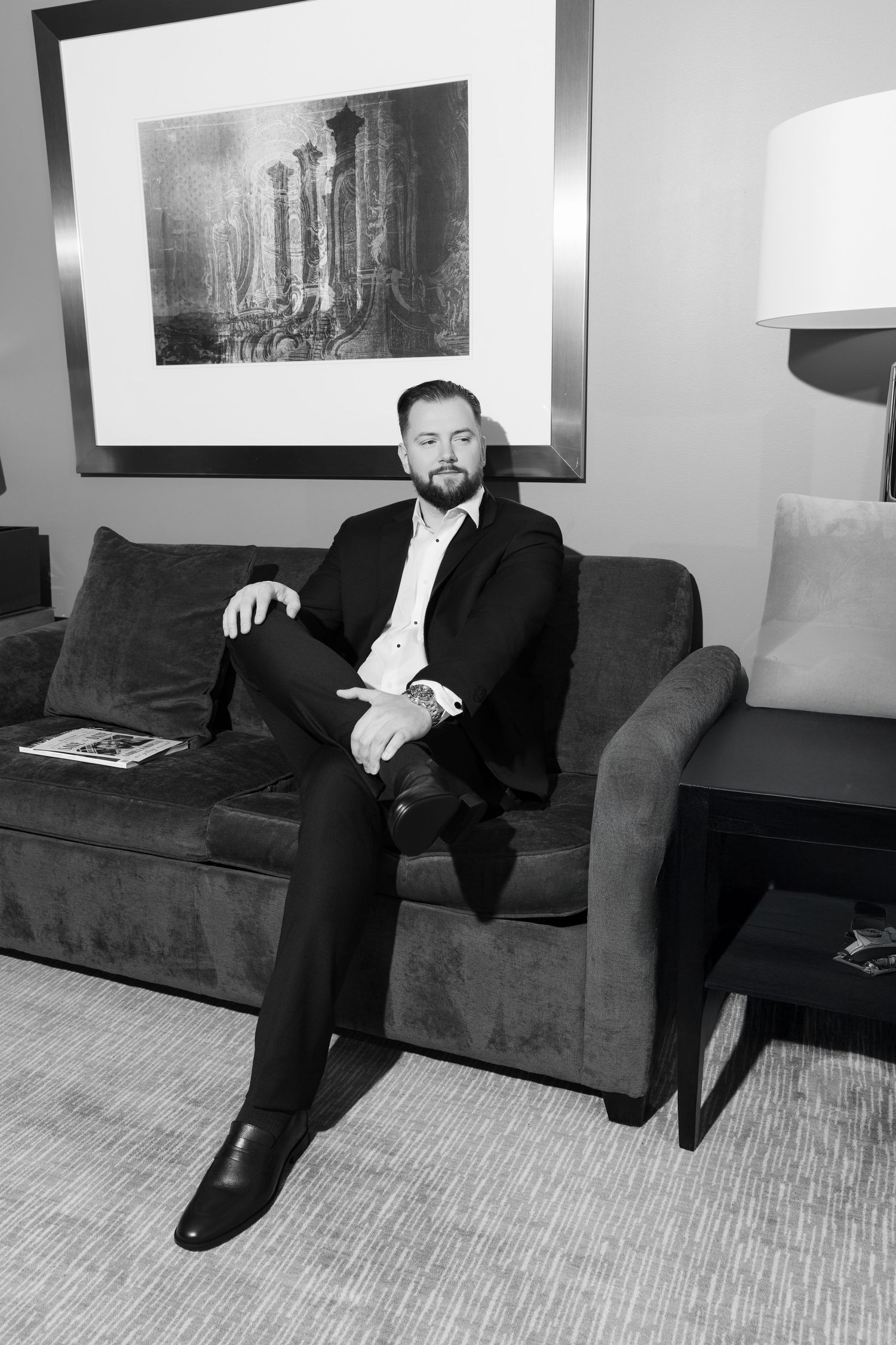 Groom in black suit sitting on gray sofa in hotel room, black and white portrait photo.