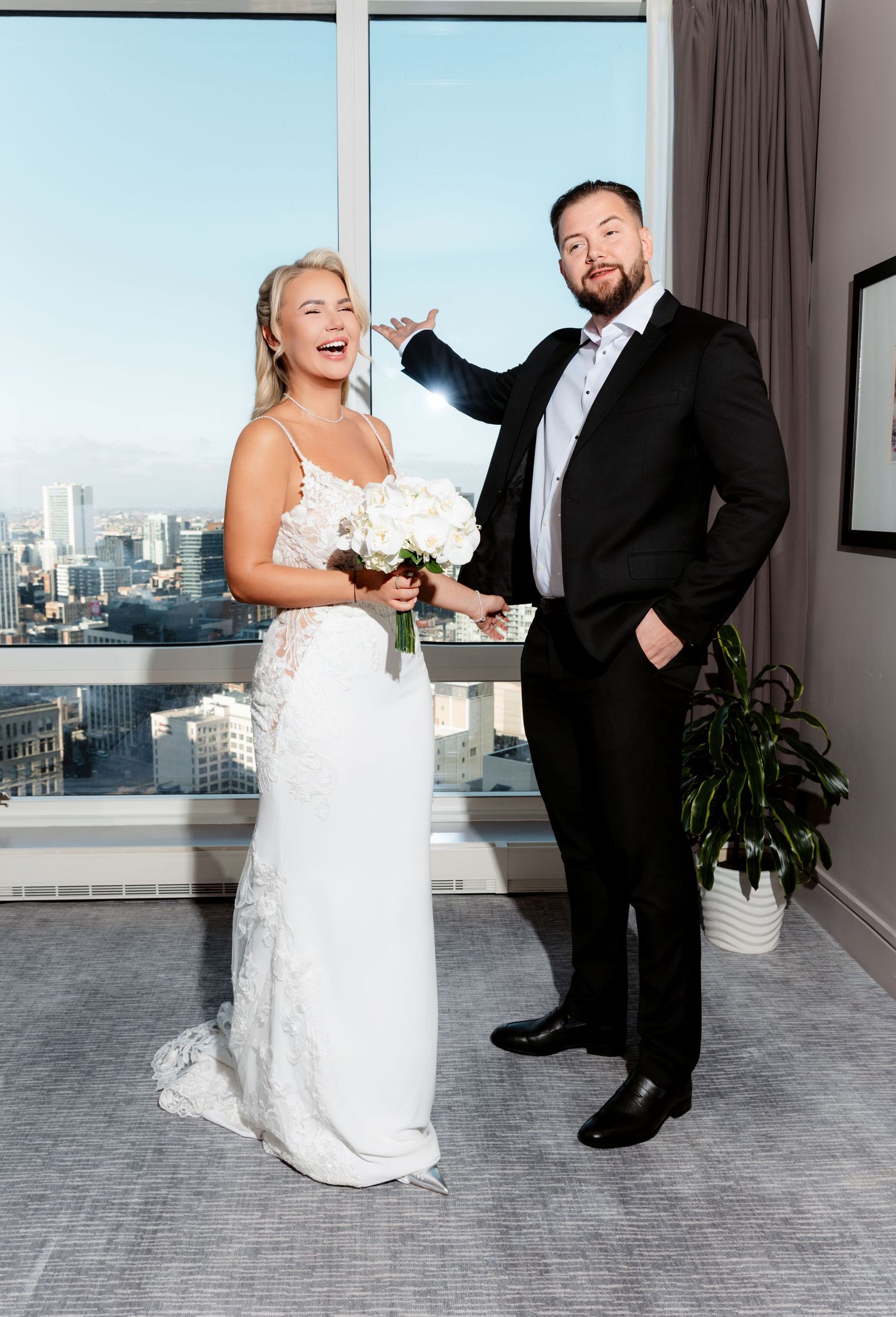 Laughing bride and groom pose playfully together in hotel suite with city skyline backdrop.