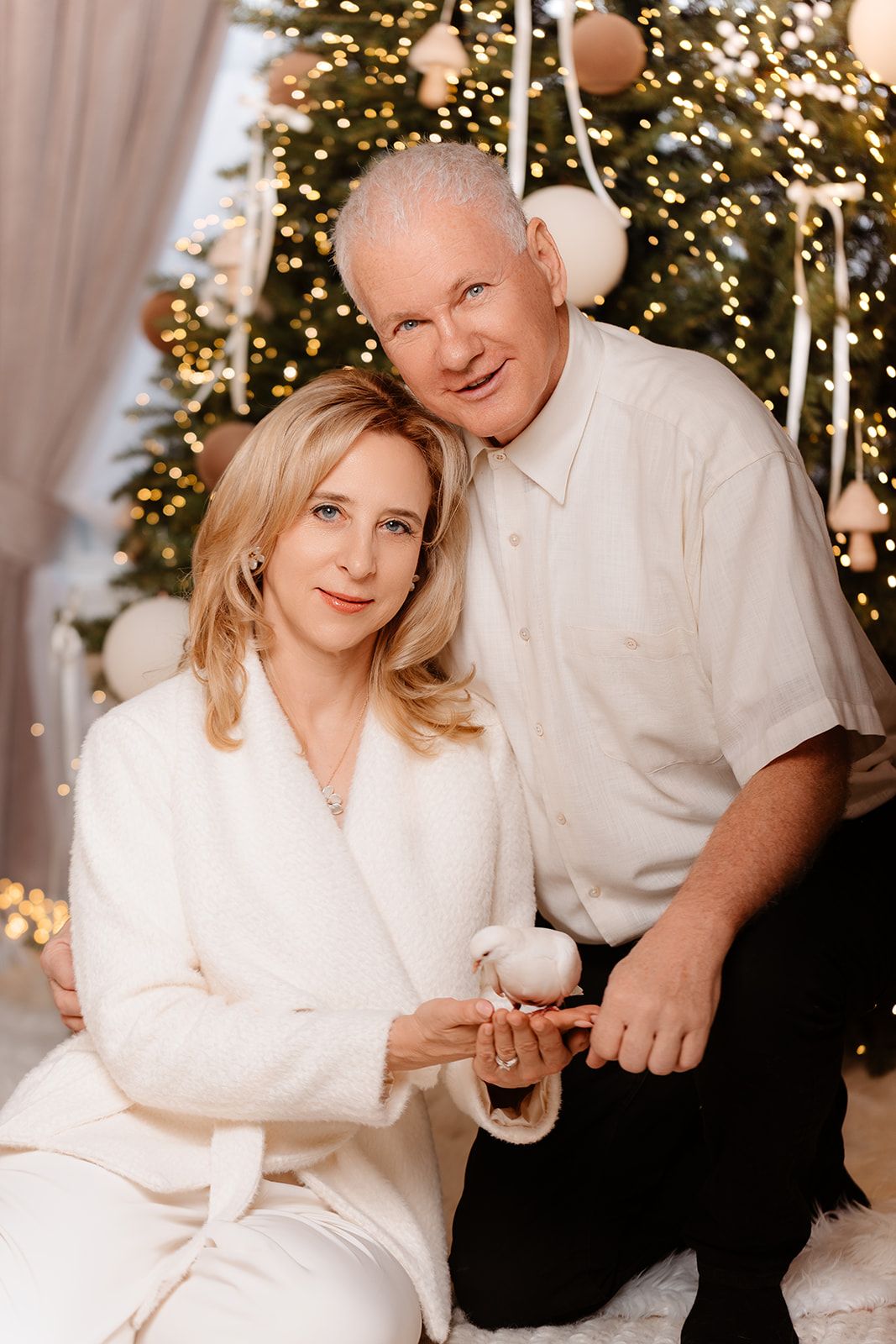 Couple in white attire posing together in front of decorated Christmas tree with warm golden lights.