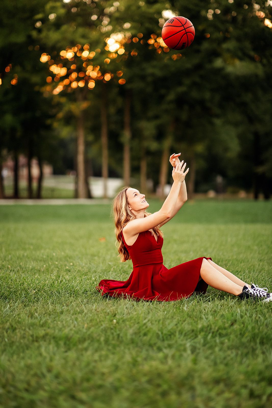 Blonde girl in red dress sitting on grass tossing a basketball in the air outdoors.