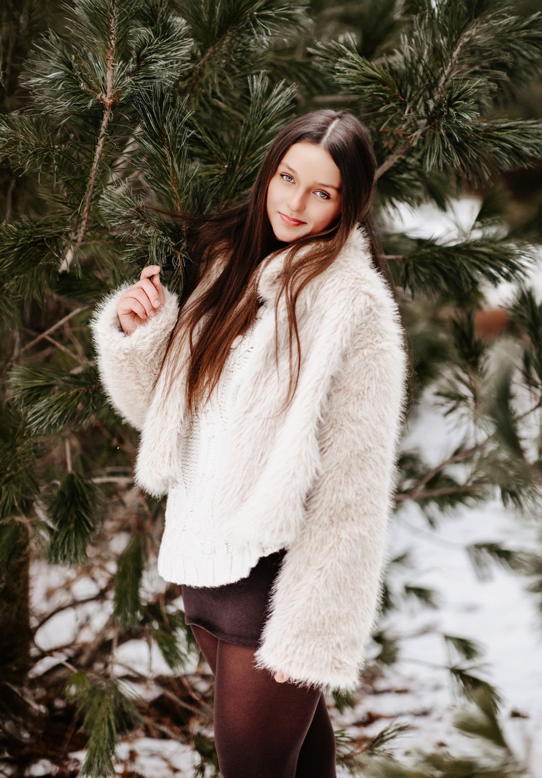 Young woman in white fluffy coat standing among snow-dusted pine trees in a winter forest setting.