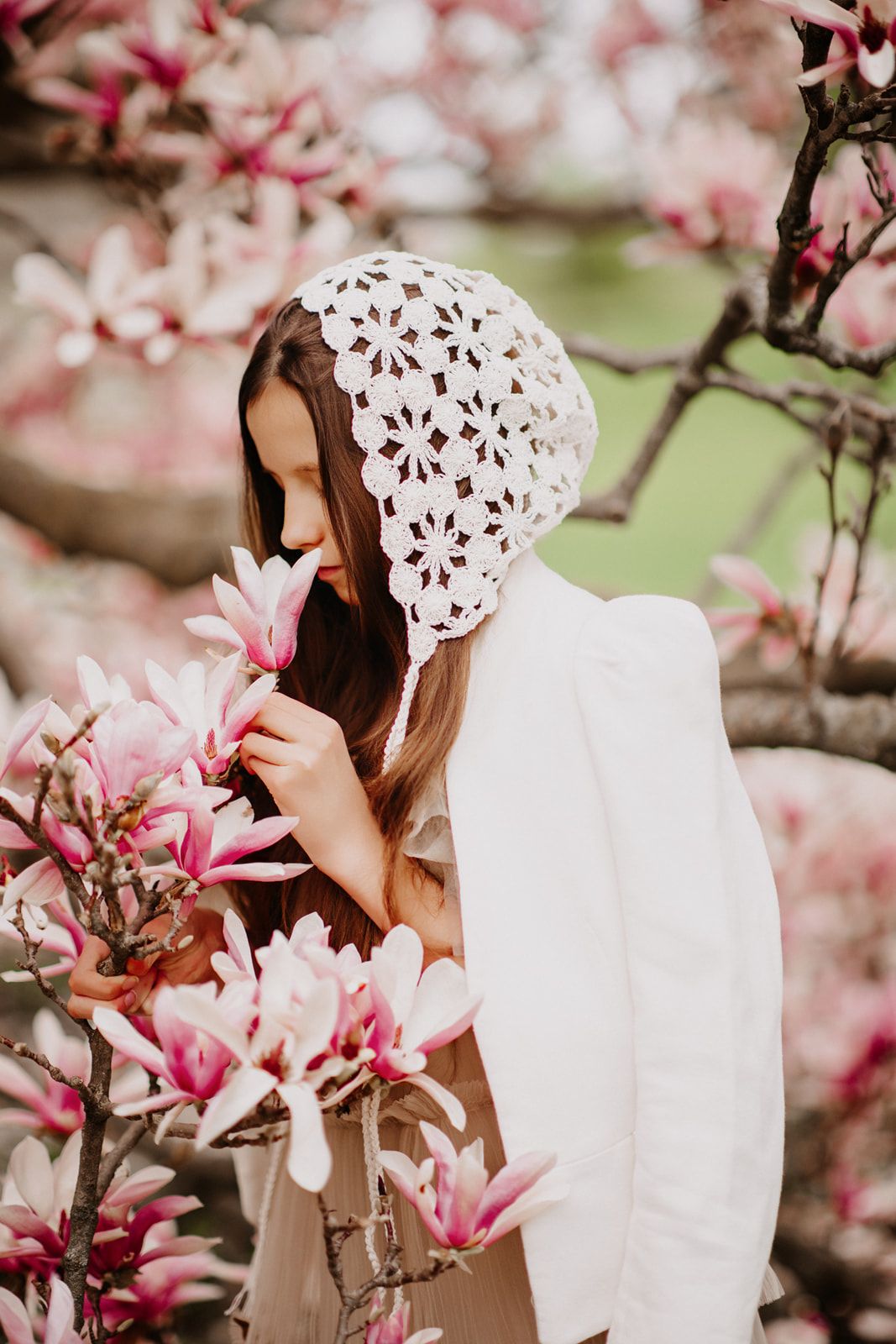 Woman in white crochet bonnet smelling pink magnolia blossoms on a flowering tree in spring.