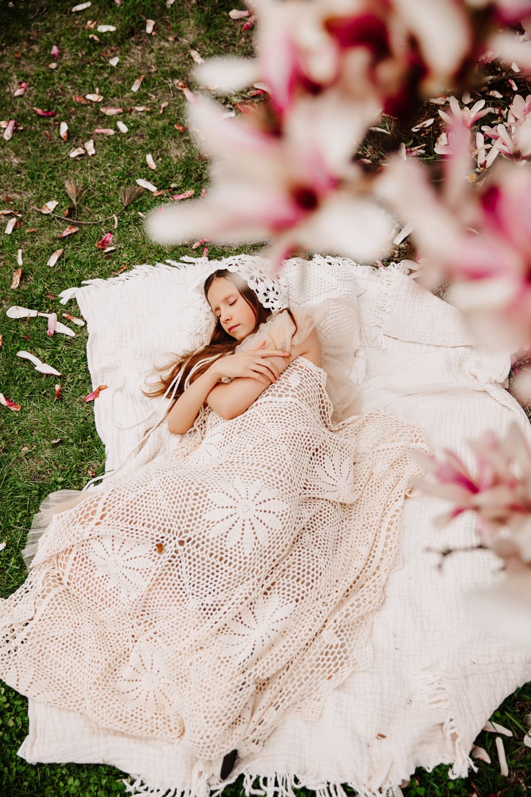 Young girl sleeping outdoors on white crochet blanket surrounded by pink magnolia petals on green grass.