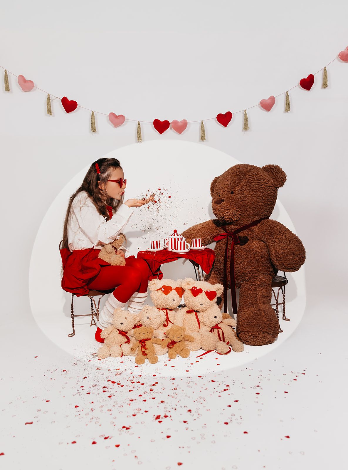 Girl blows confetti at giant teddy bear during Valentine's Day tea party with small bears and heart garland decor.