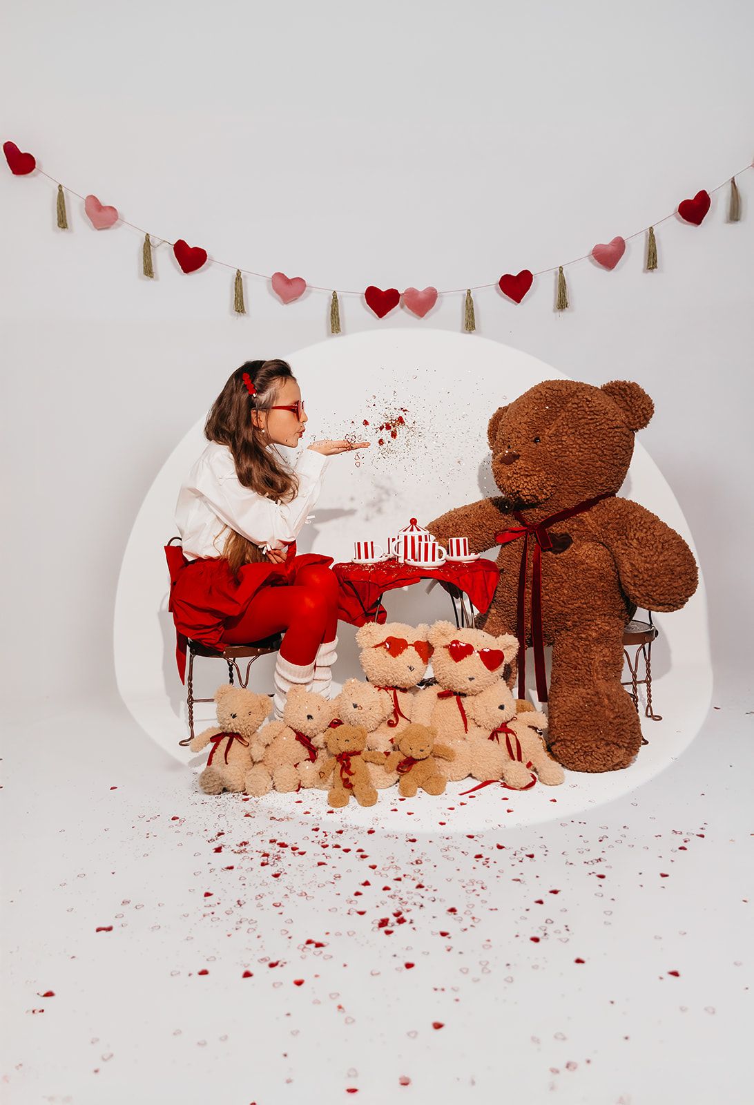 Girl having a Valentine's tea party with large brown teddy bears, red decor, heart garland, and confetti in a white studio.