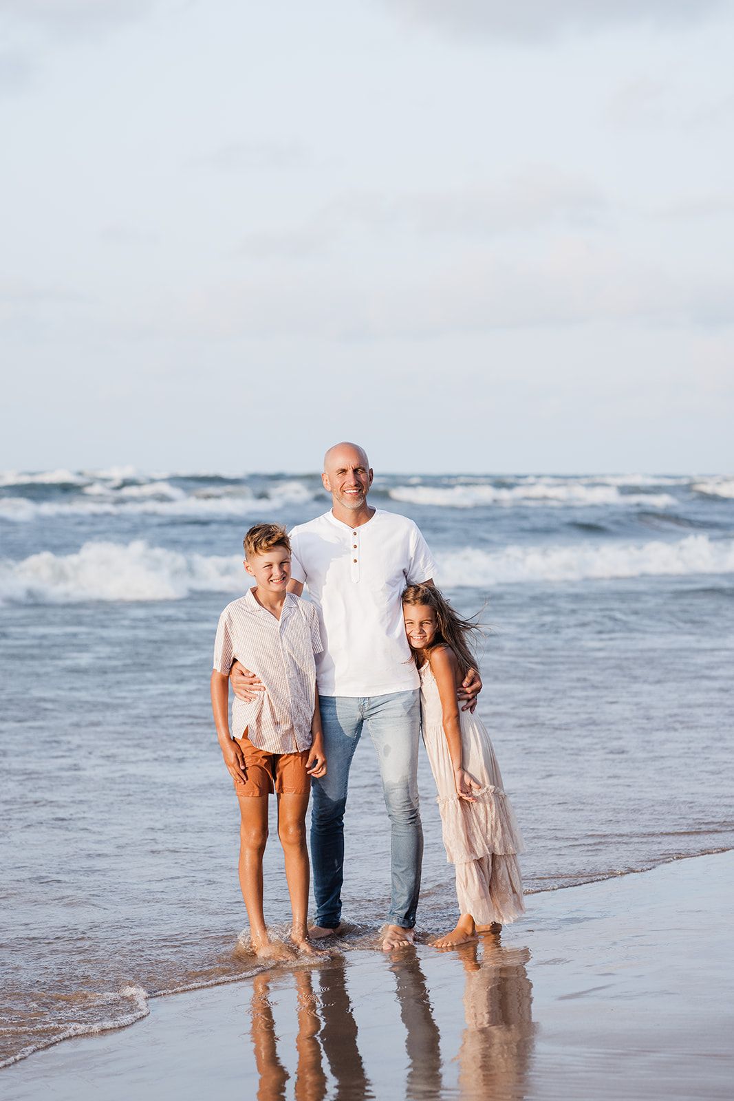 A father poses with his two children on a sunny beach, waves crashing behind them, all wearing white outfits.