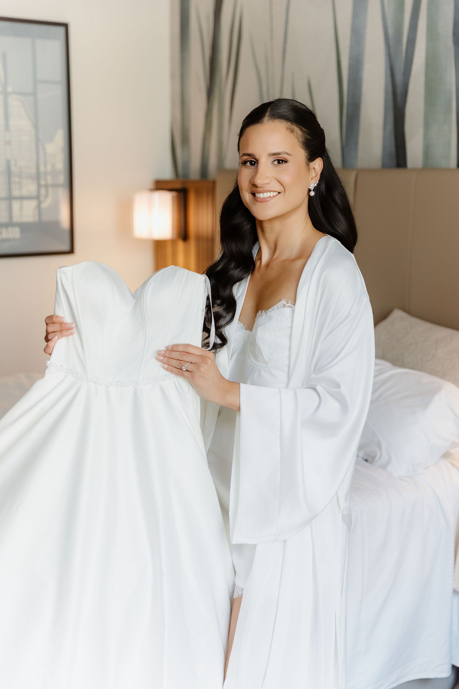 Smiling dark-haired bride in white robe holds strapless wedding gown in bright hotel room before getting dressed.