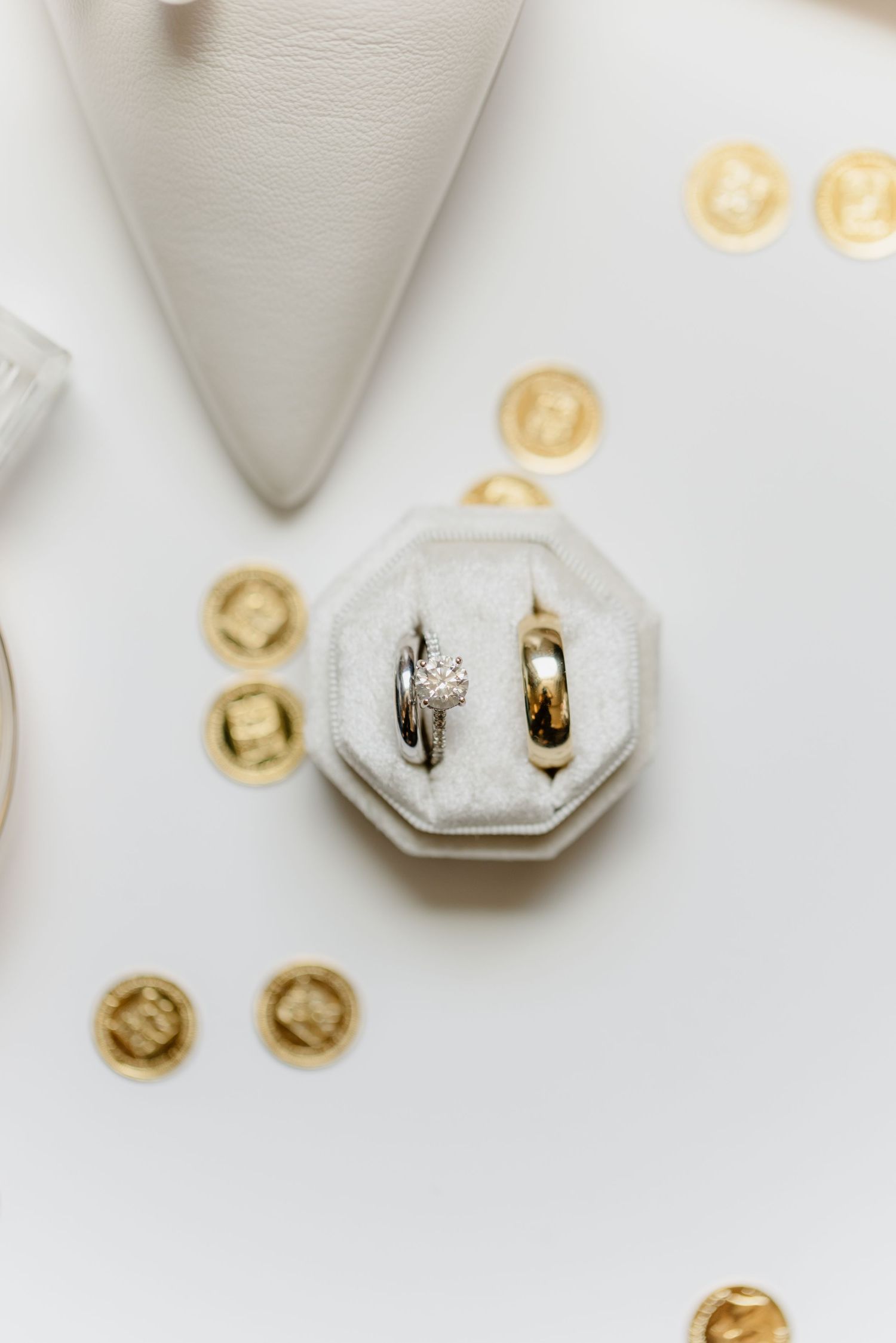 Diamond engagement ring and gold wedding bands displayed in white velvet octagonal ring box with gold coins.