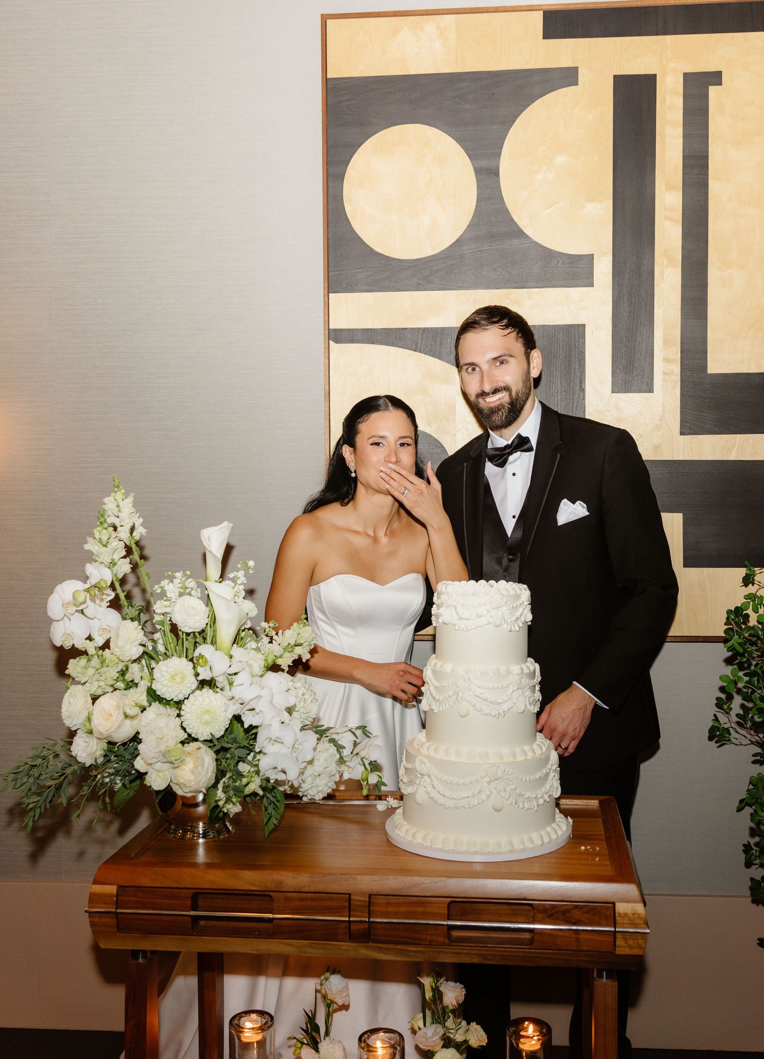Bride covering her mouth in surprise while groom smiles beside elegant white tiered wedding cake.