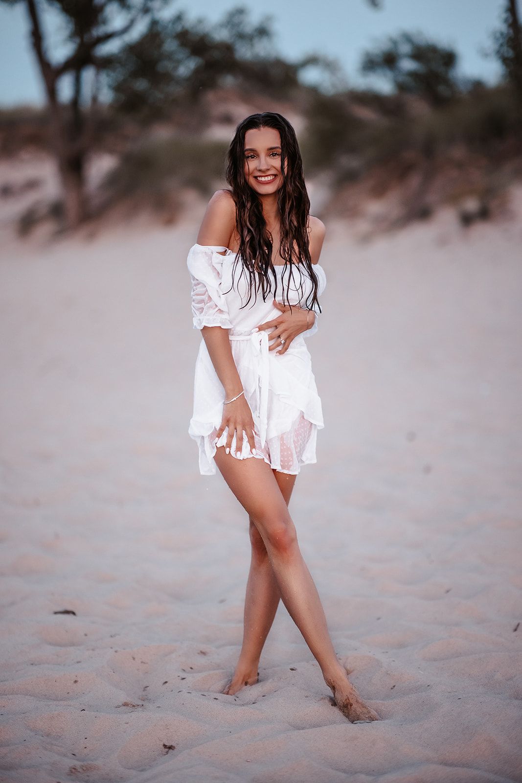 Smiling woman in white off-shoulder dress posing barefoot on sandy beach at dusk.