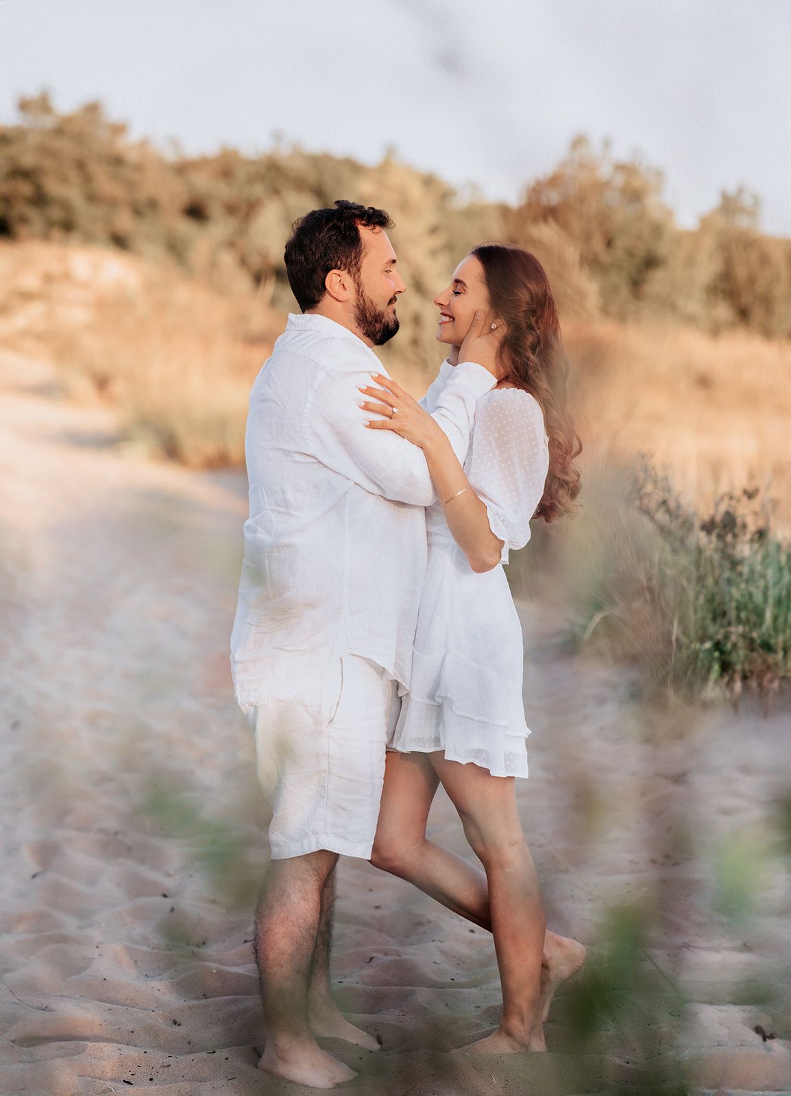 A couple in matching white outfits embrace and smile at each other on a sandy beach path.