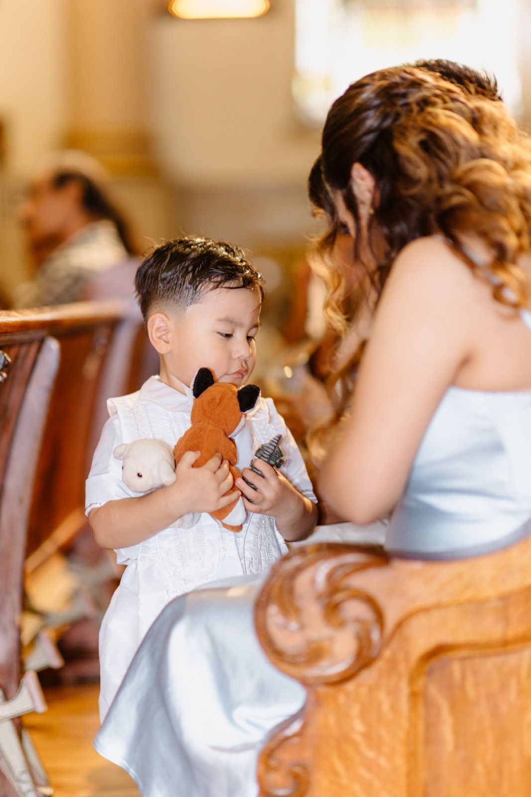 Young boy in white outfit holds stuffed animals beside a woman in a church pew during a baptism ceremony.