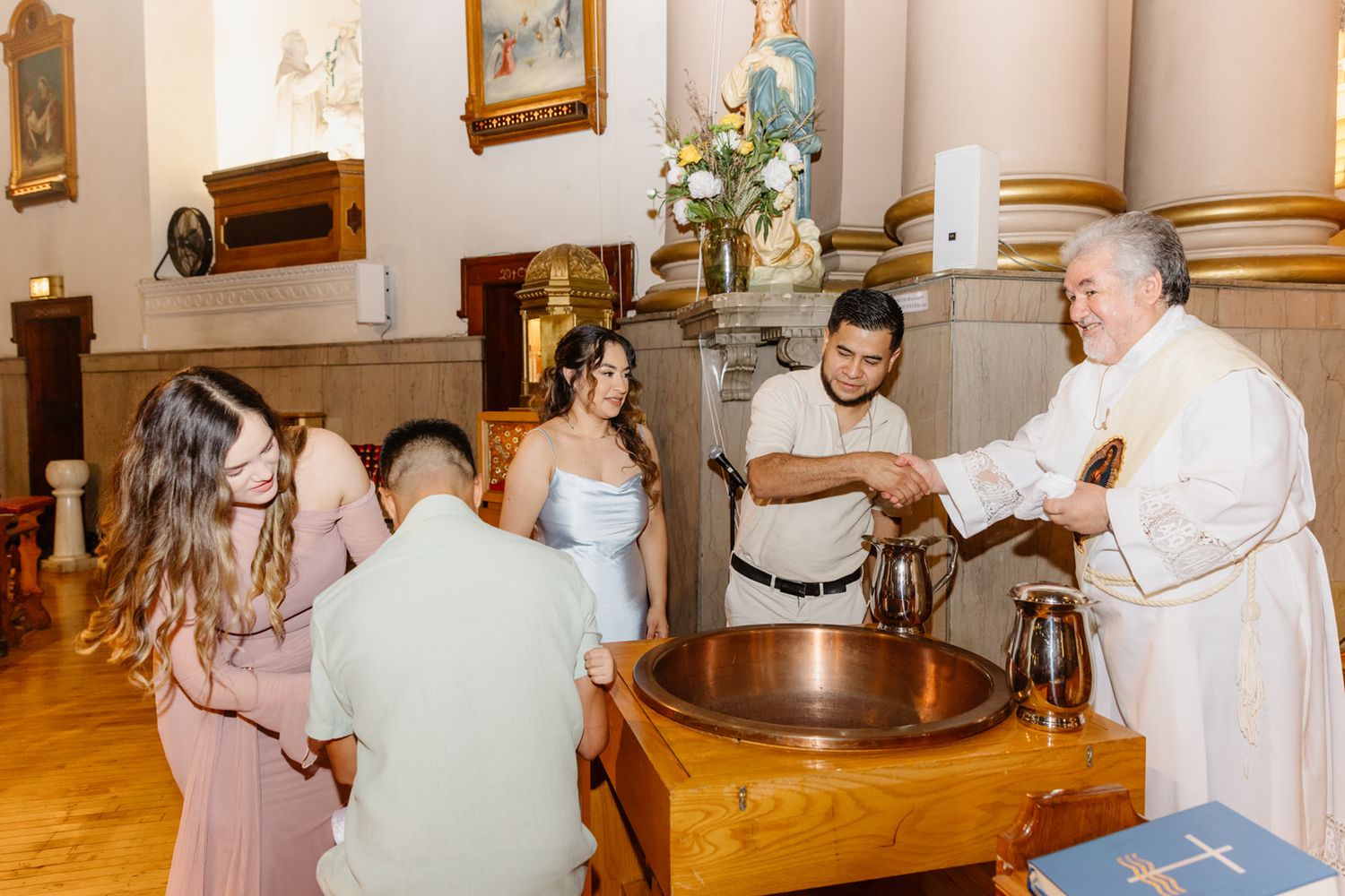 Priest performs baptism ceremony over copper font with family gathered in a historic Catholic church.
