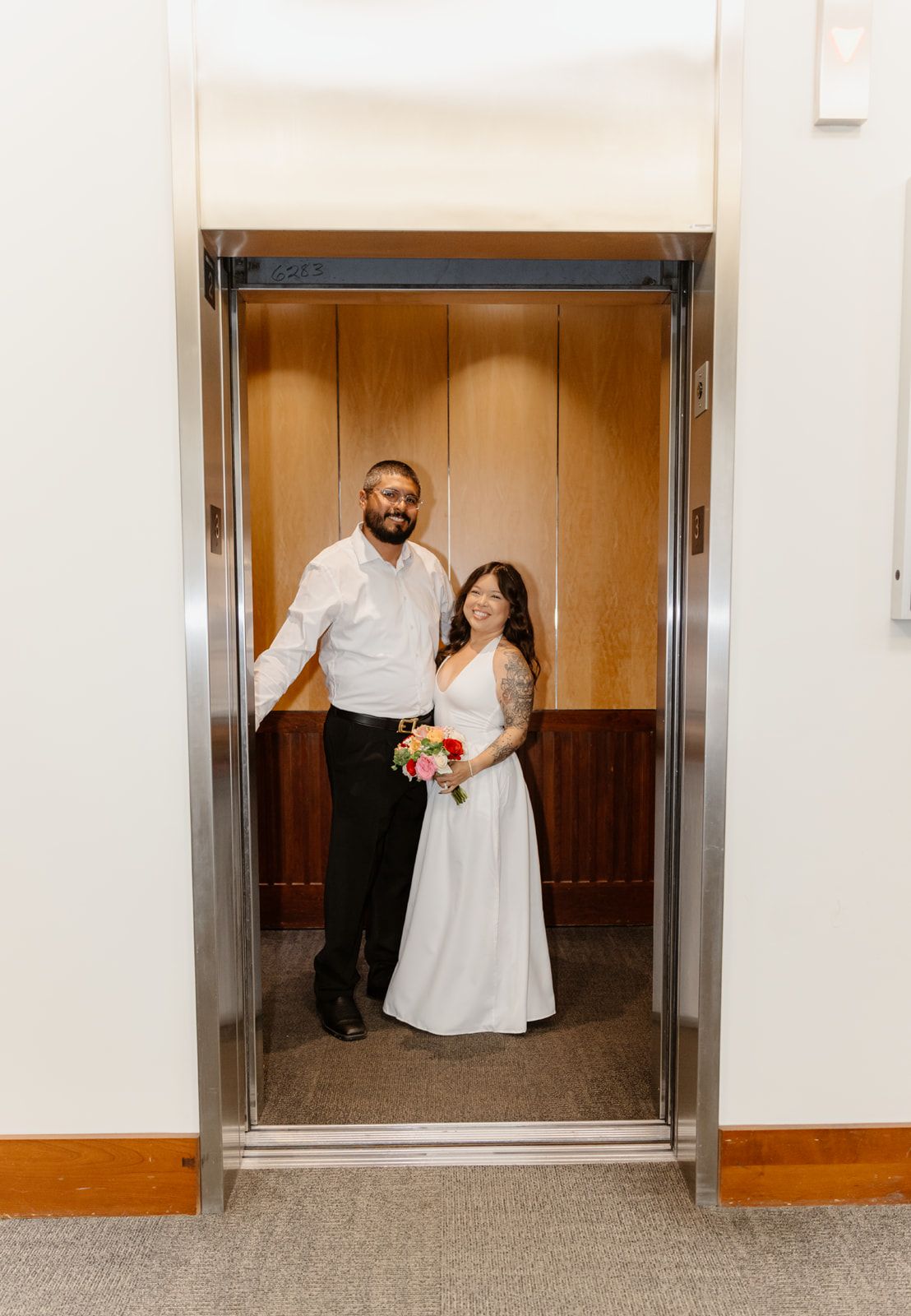 Newlywed couple smiling in elevator, bride in white gown holding colorful bouquet, groom in white shirt and black pants.
