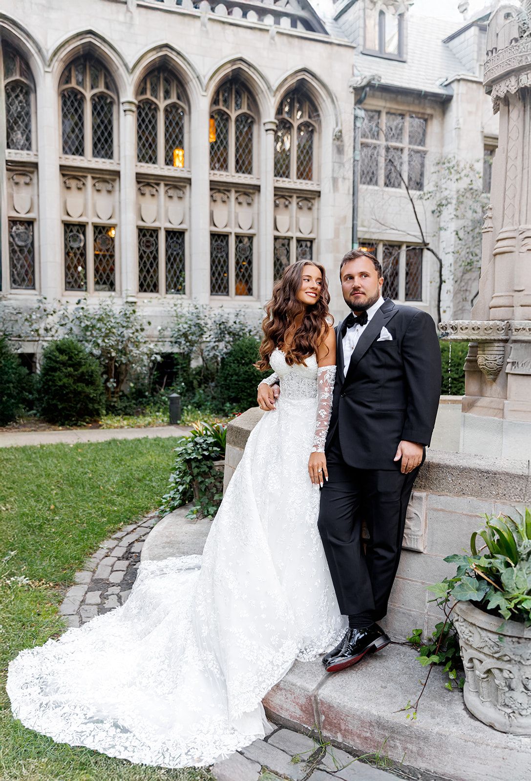 Elegant bride in lace gown and groom in tuxedo pose together in a Gothic church courtyard garden.