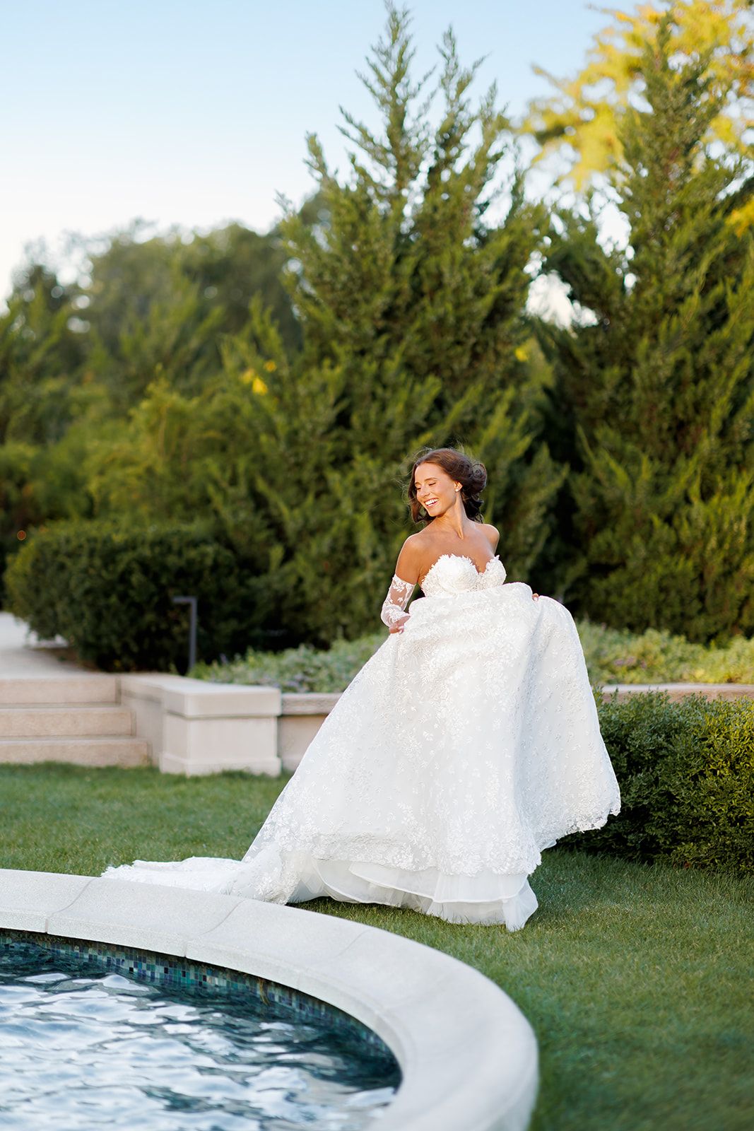 Smiling bride in strapless lace ballgown lifts her skirt near a fountain surrounded by tall evergreens.