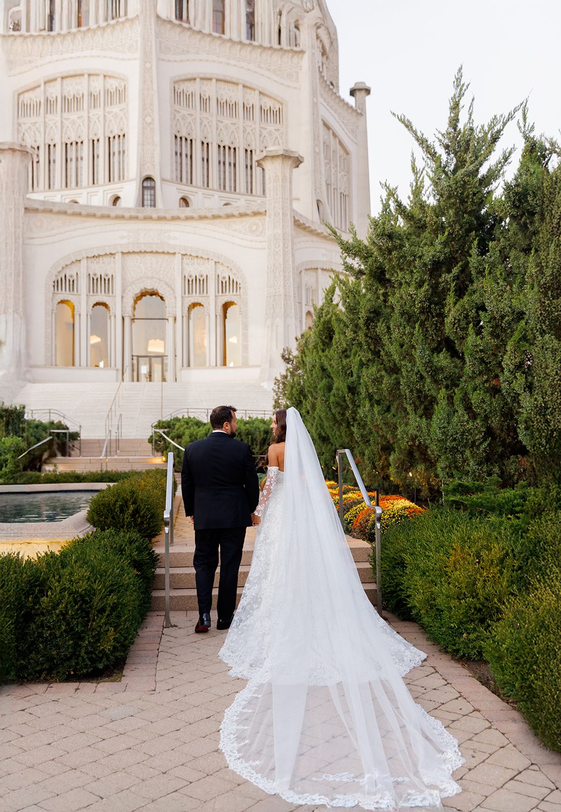 Bride and groom walk hand-in-hand toward a grand white temple surrounded by lush greenery, bride's lace veil trailing elegantly.