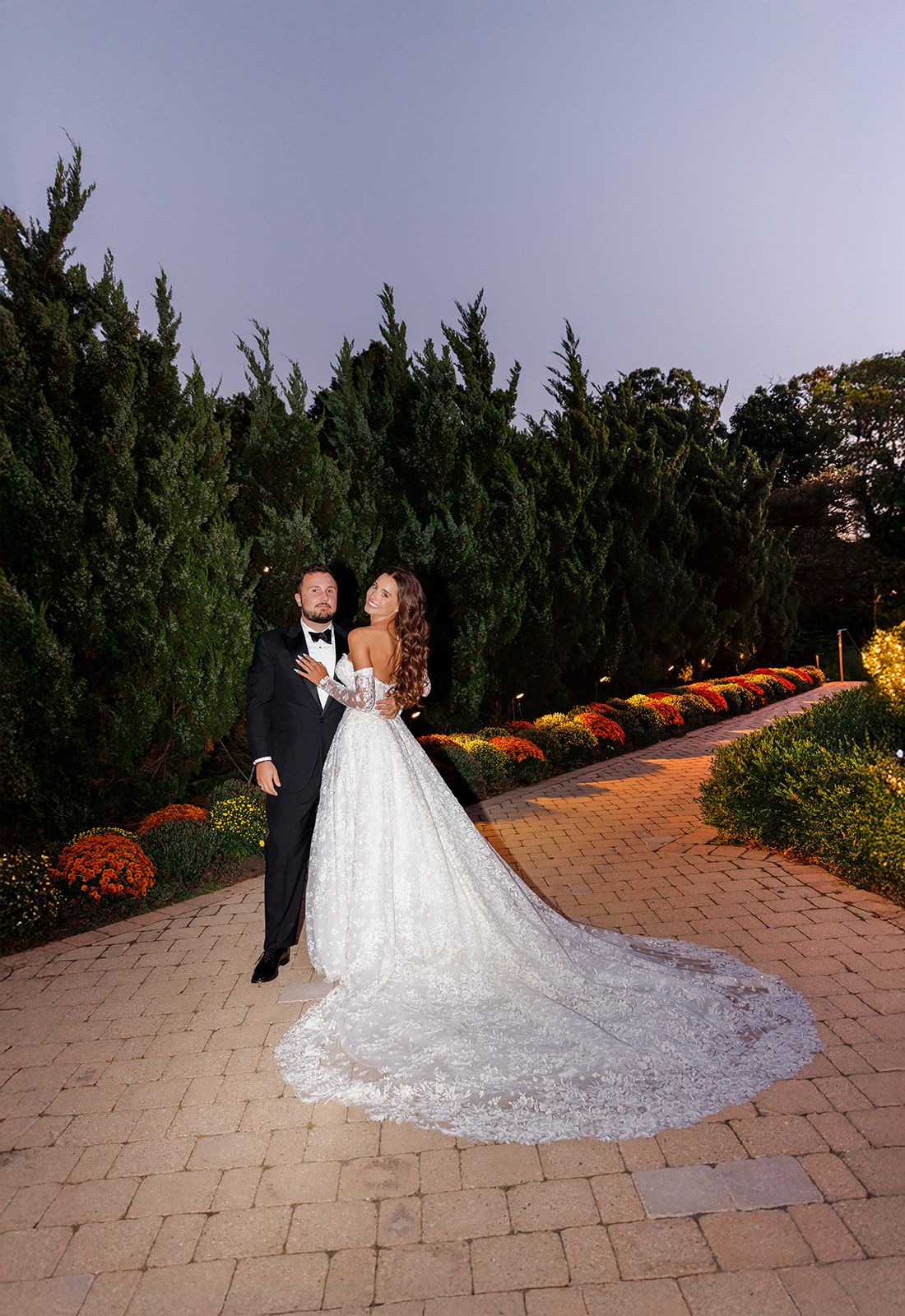 Bride in lace ball gown and groom in tuxedo pose on brick path surrounded by tall evergreens and colorful fall mums at dusk.