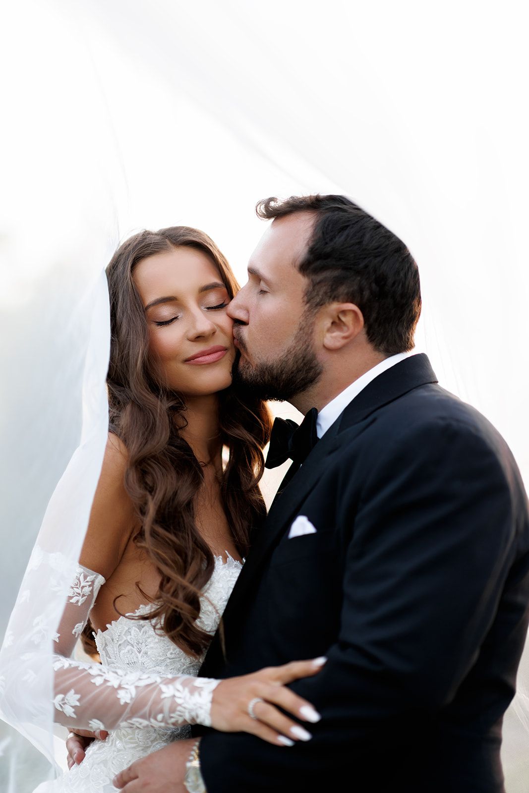 Groom in black tuxedo kissing bride on cheek as she smiles with eyes closed under wedding veil.