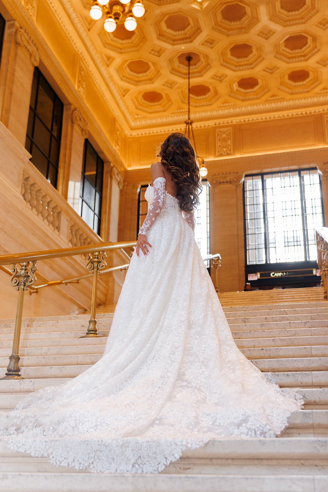Bride in elegant lace wedding gown with long train ascending grand marble staircase in ornate golden ballroom.