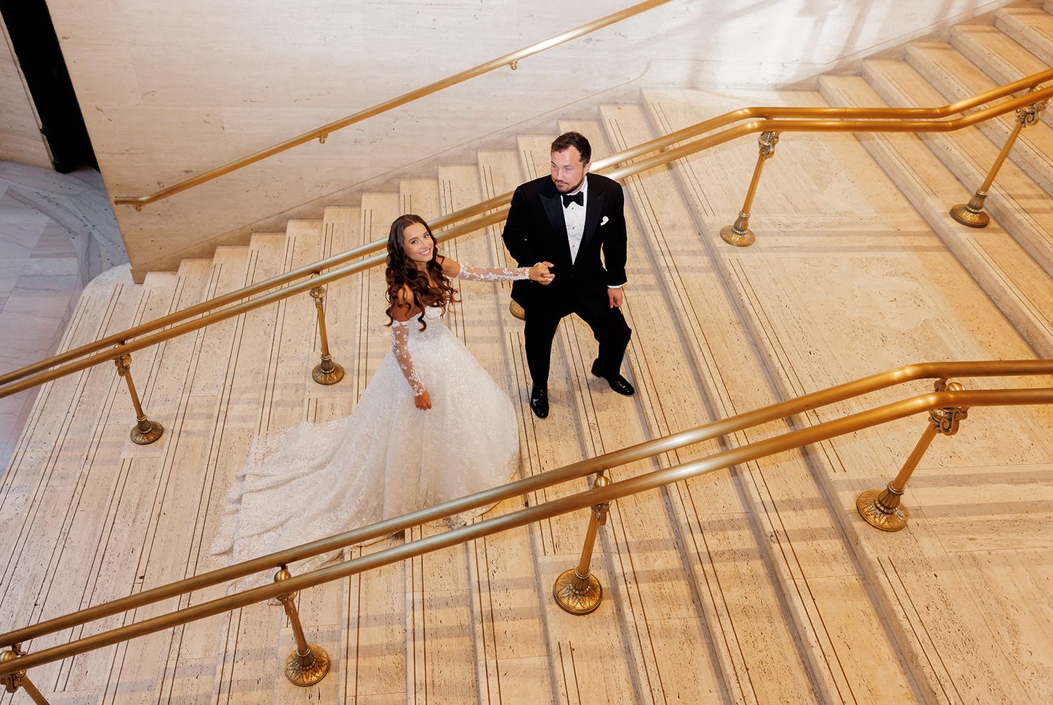 Bride and groom walking up grand staircase with gold railings, aerial view of wedding couple in formal attire.