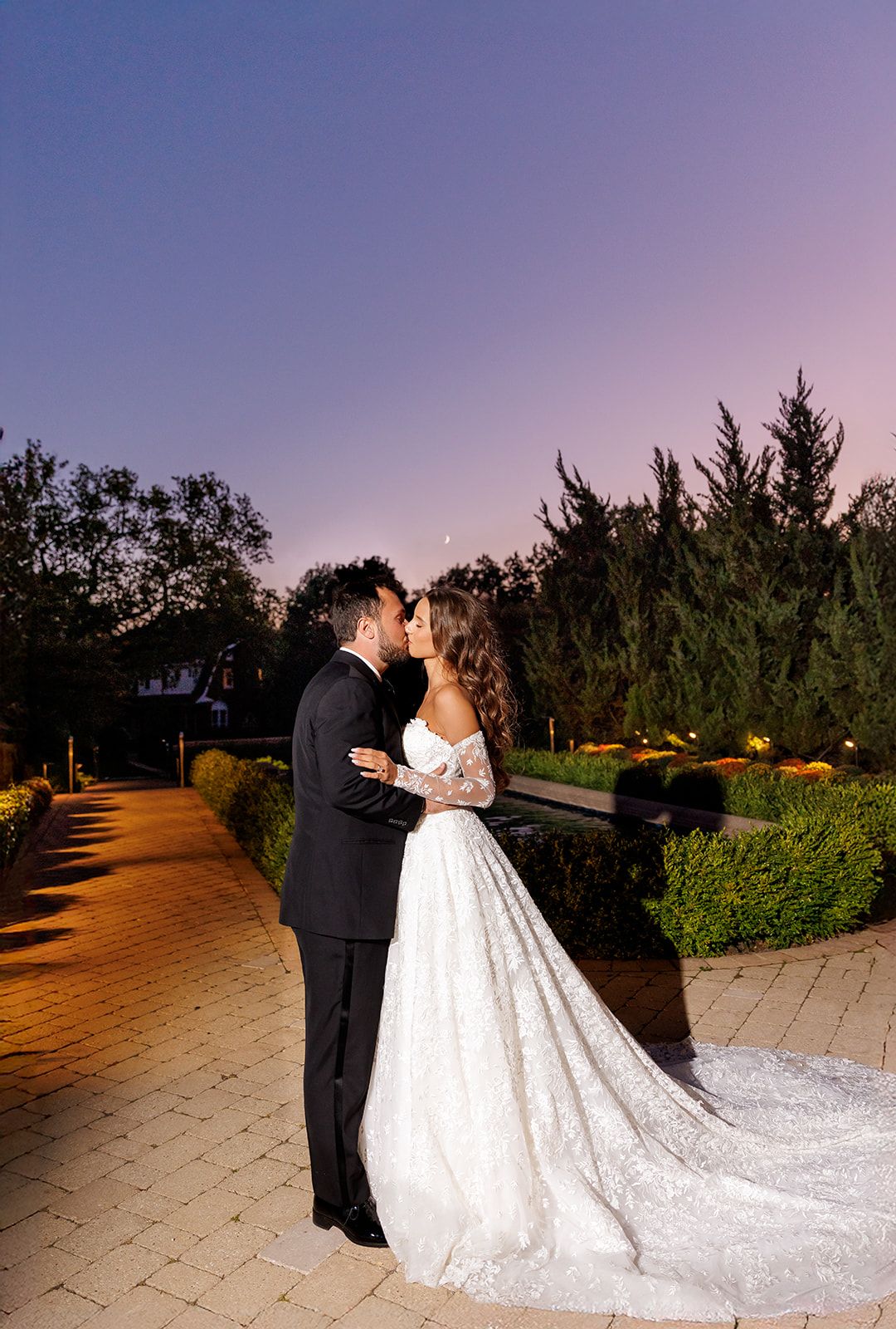 Bride and groom share a romantic kiss at dusk in a garden, bride wearing a lace ball gown with long train.