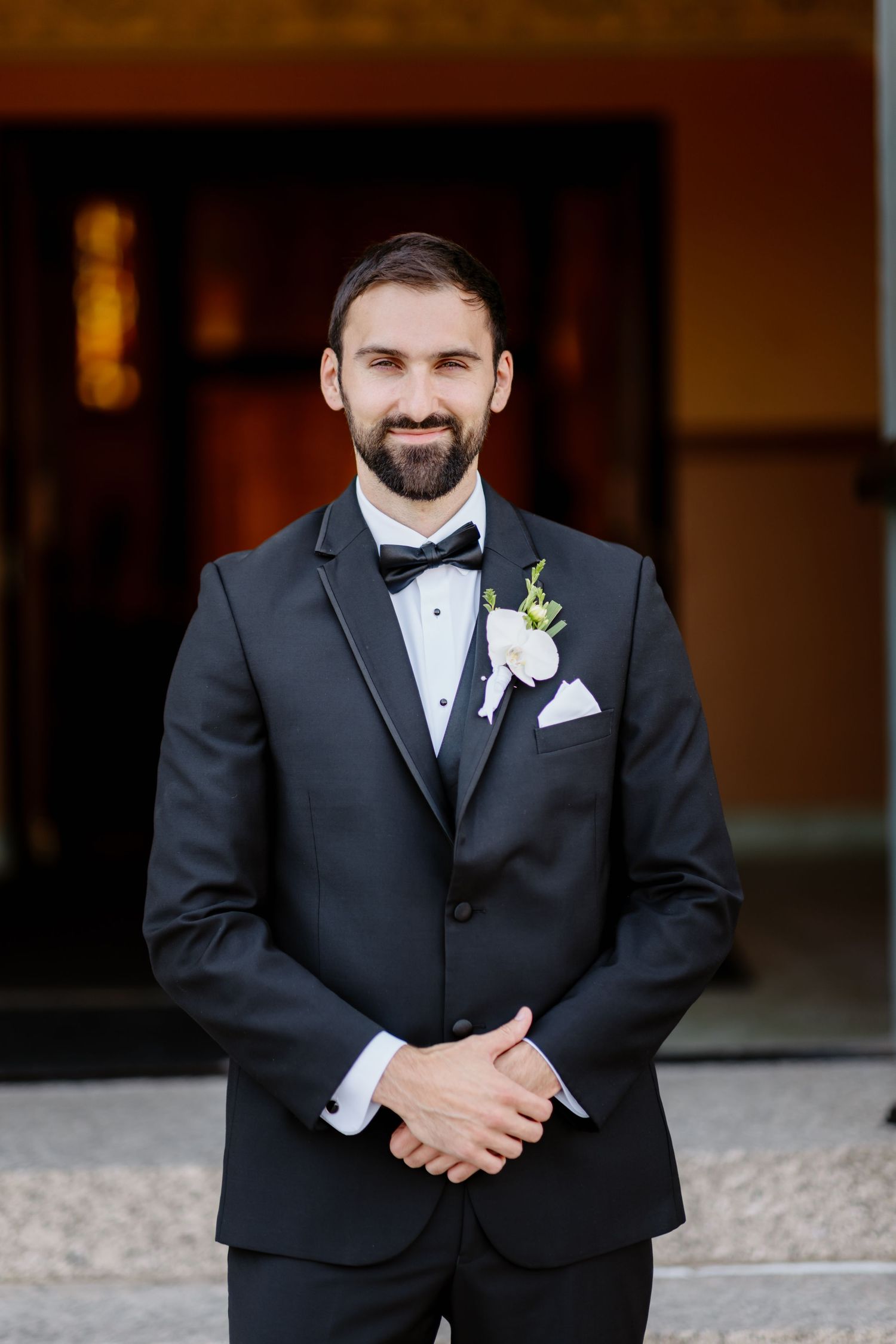 Smiling groom in black tuxedo with bow tie and white boutonniere stands confidently at wedding venue entrance.