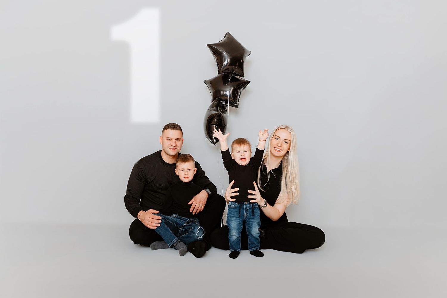 Family of four sitting on floor with black star balloons and number one for a birthday photo.