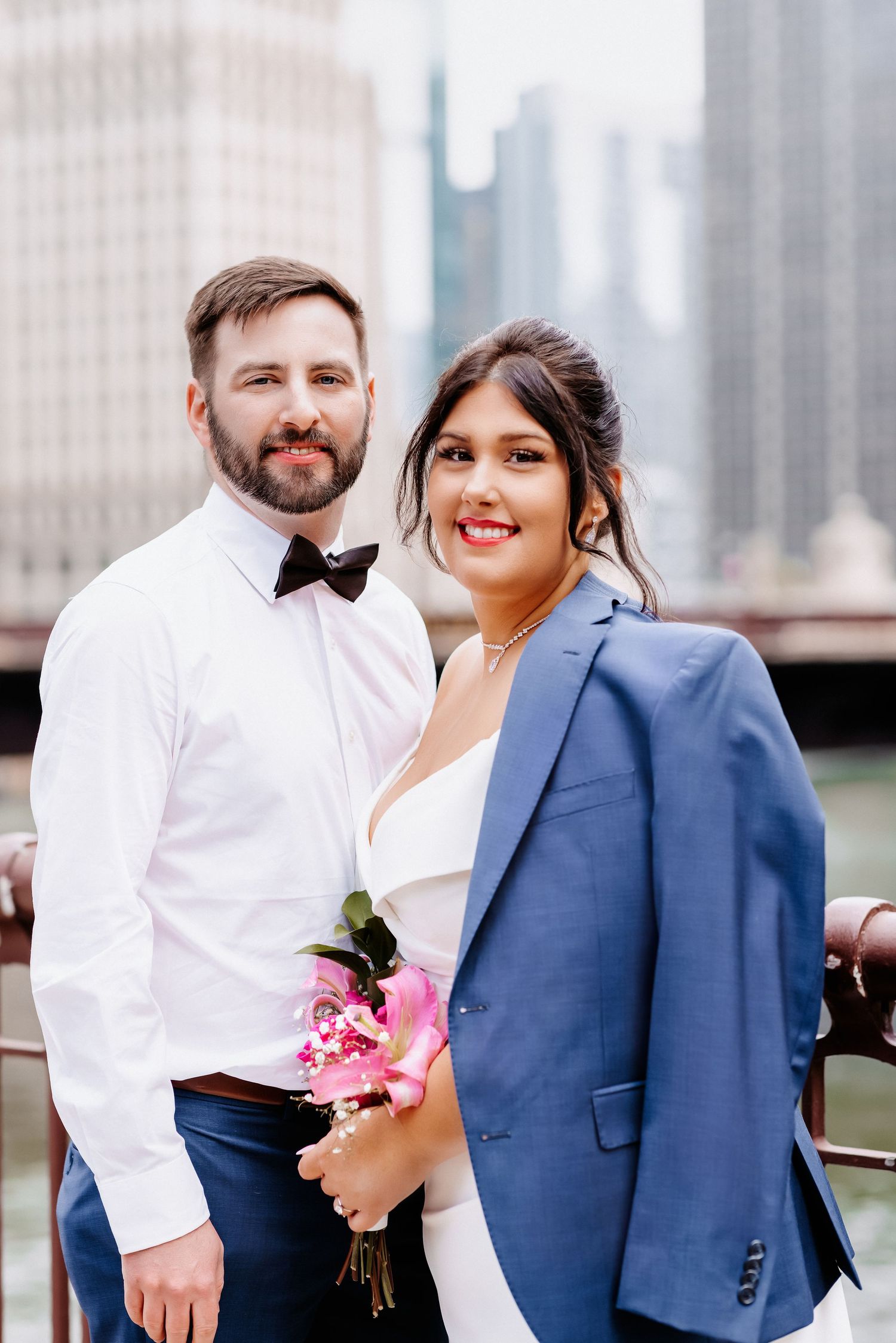 Smiling wedding couple poses on Chicago riverfront bridge, bride holding pink bouquet wearing blue blazer over gown.
