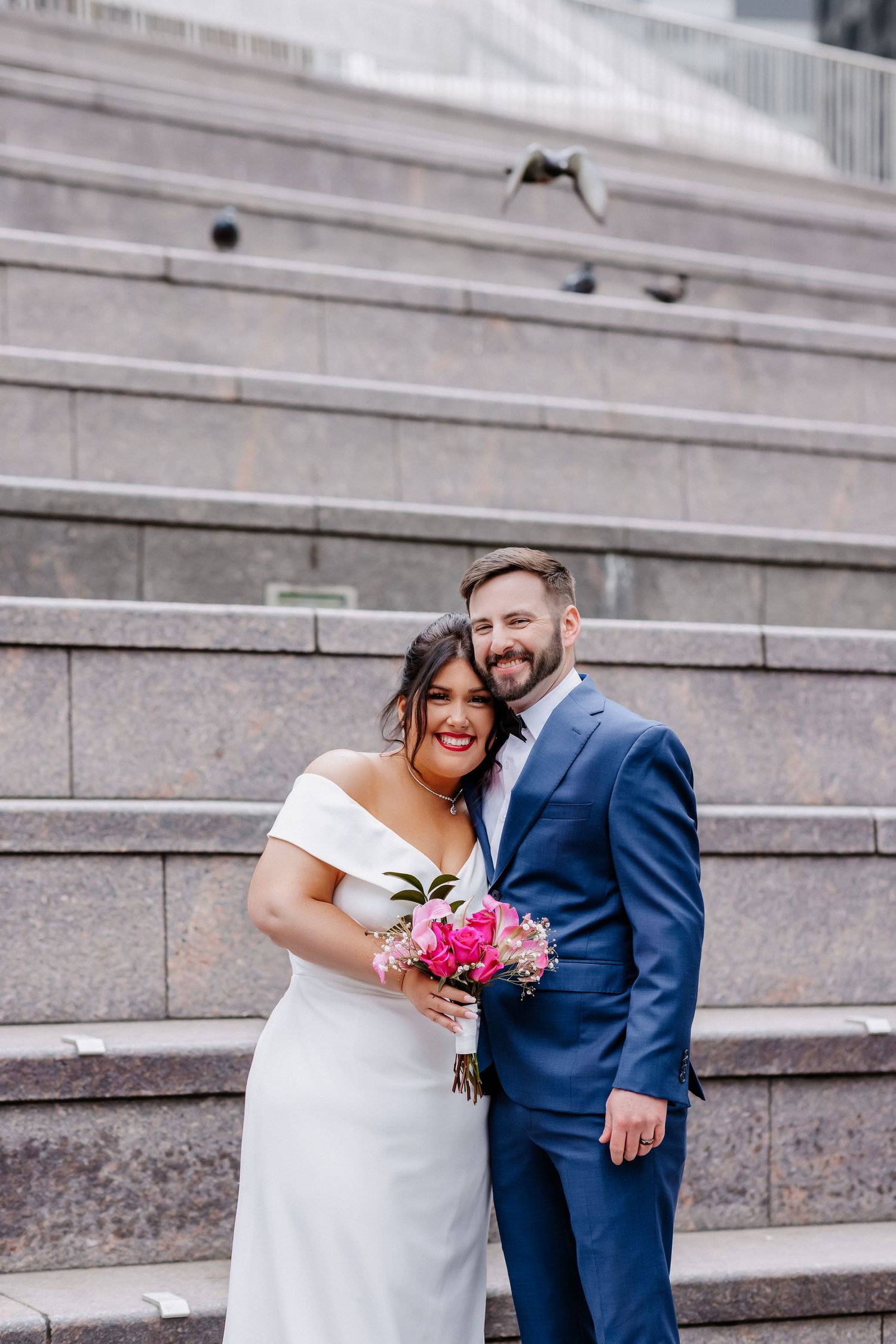 Bride and groom pose on stone steps, she holds pink bouquet, bird flies overhead in background.