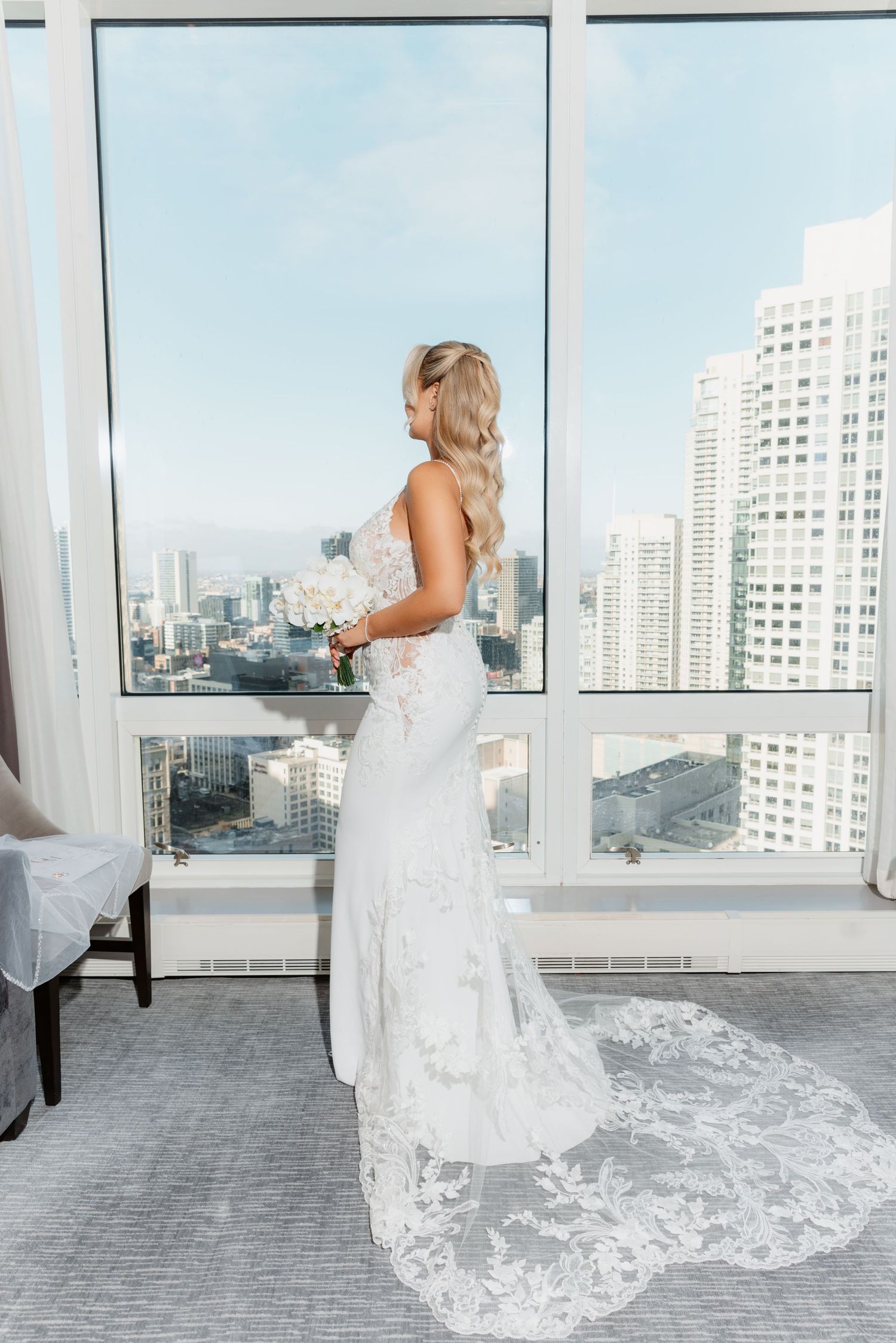 Bride in backless lace wedding dress with long train gazes at city skyline through floor-to-ceiling windows.