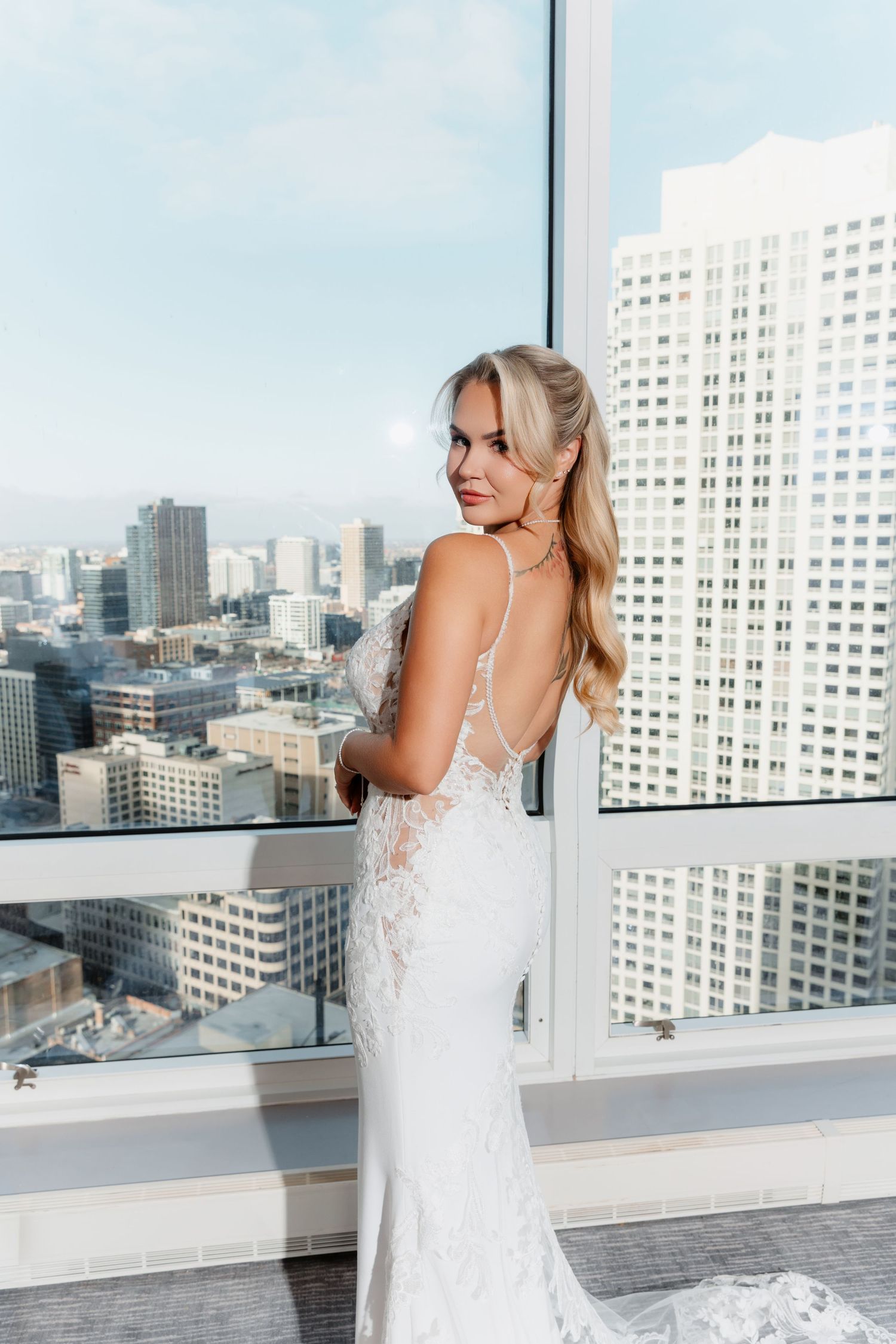 Blonde bride in elegant backless lace wedding gown smiles over shoulder with city skyline backdrop.