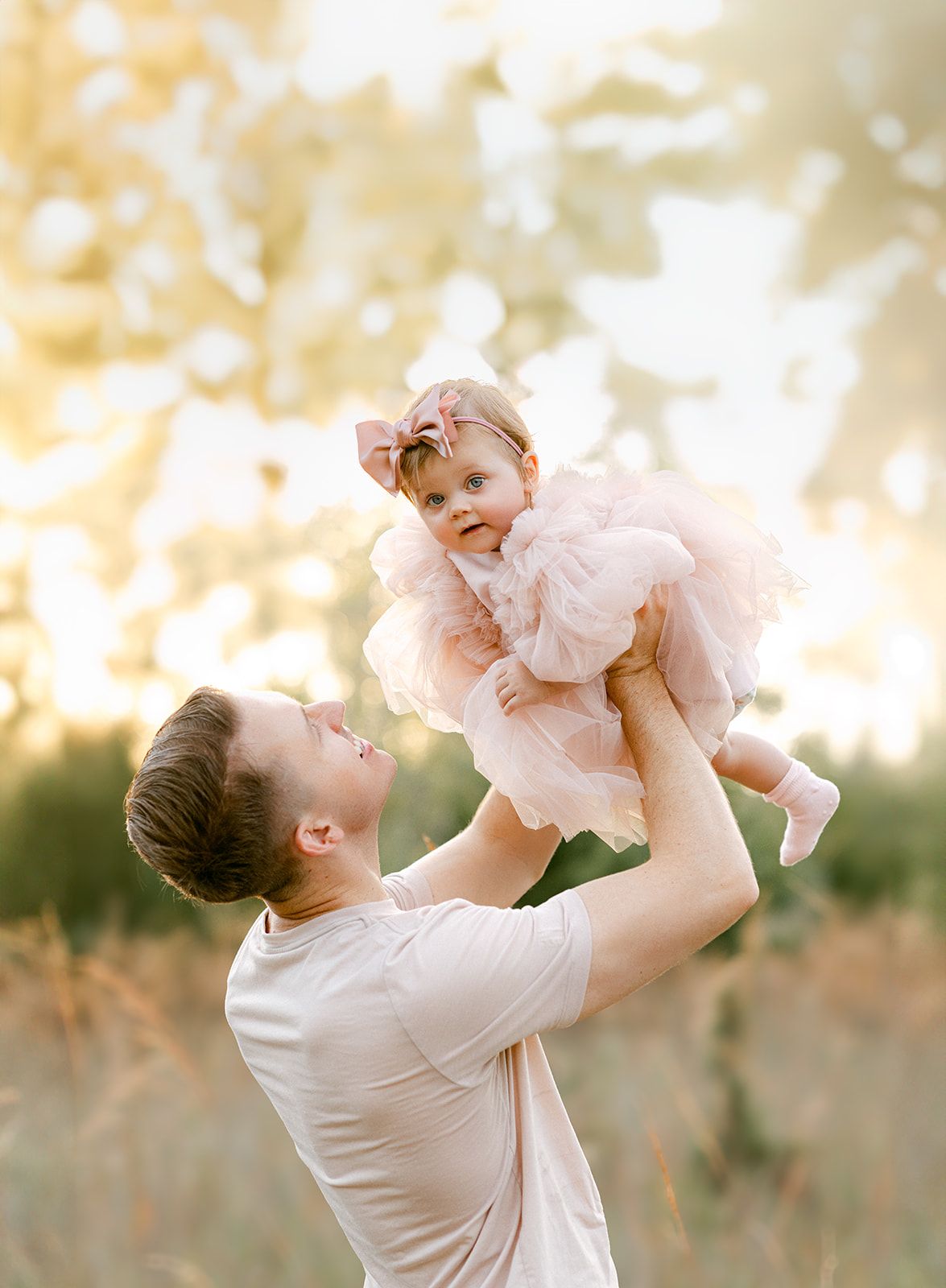 Father lifts joyful baby girl in pink tulle dress high in the air in a sunlit meadow.