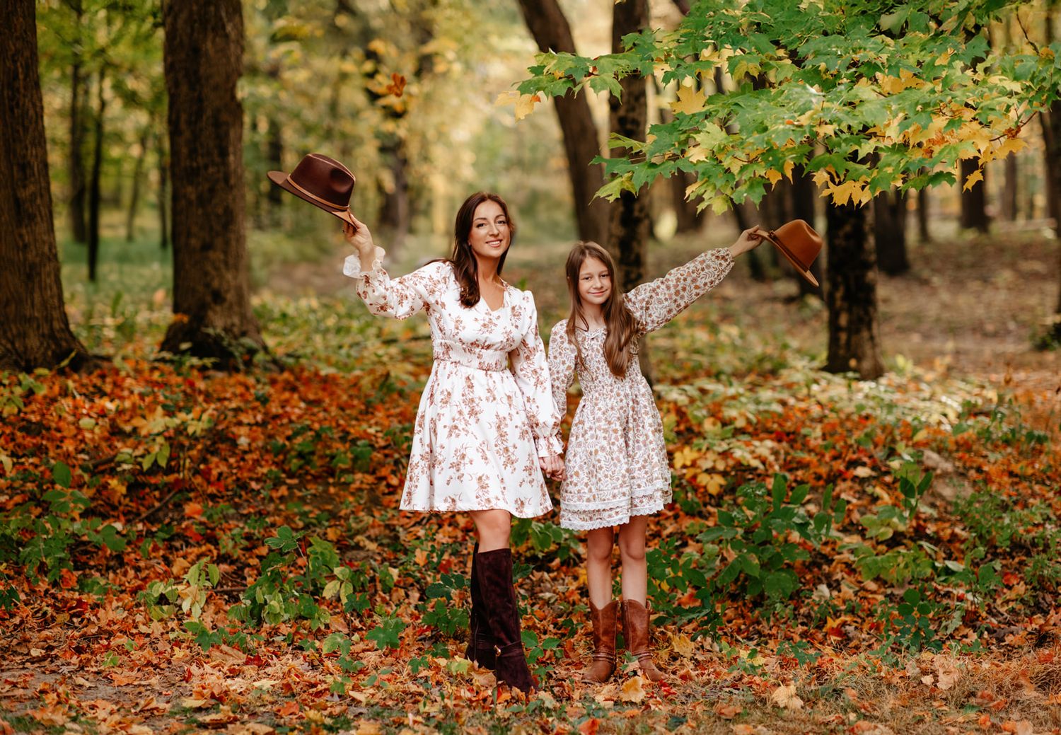 Mother and daughter in matching floral dresses tossing autumn leaves in a colorful fall forest.