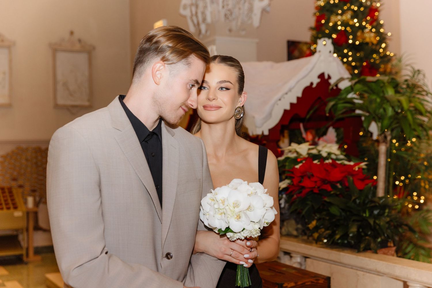 Couple in festive wedding attire posing with white bouquet near Christmas decorations.