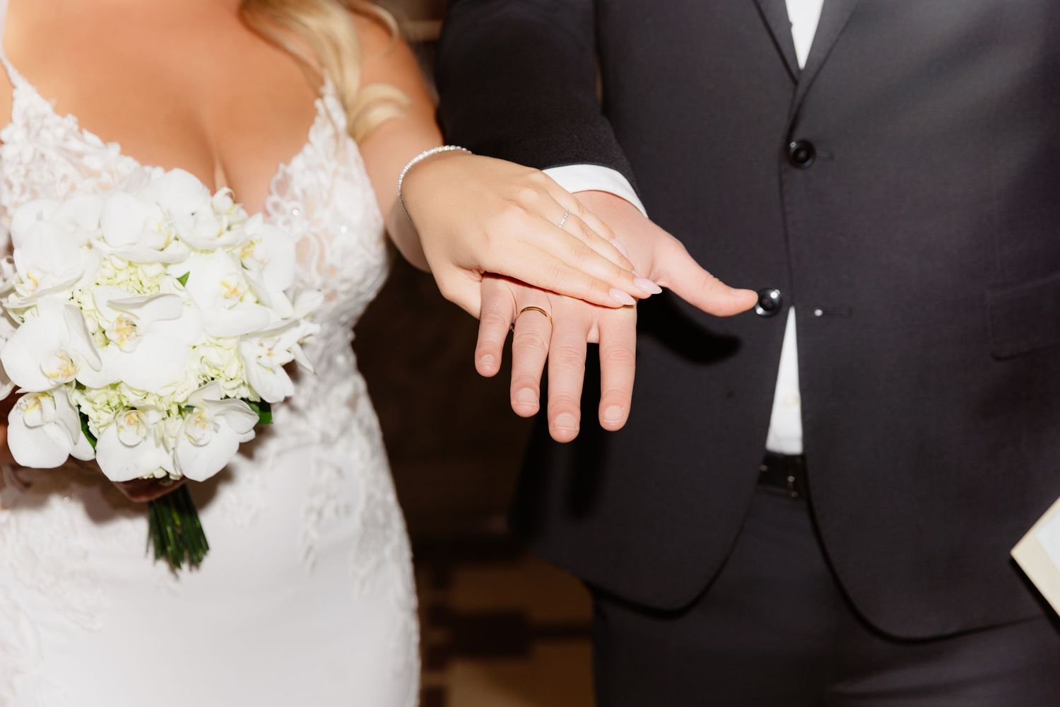 Bride and groom displaying wedding rings on their hands, bride holding white floral bouquet in lace wedding dress.