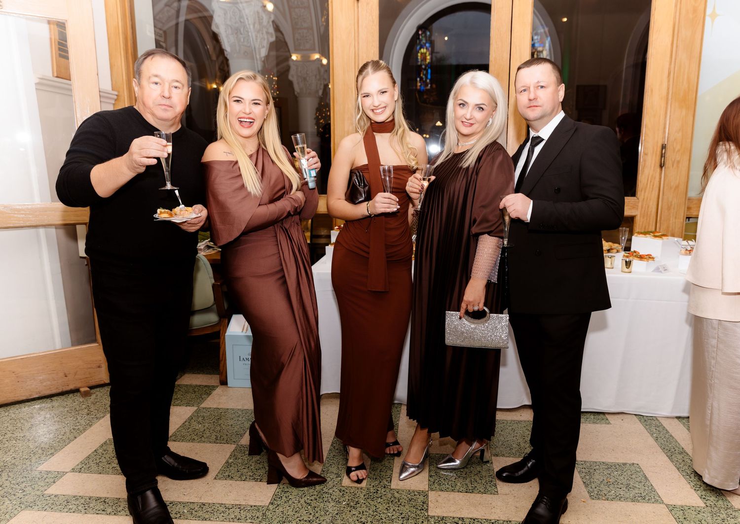 Five elegantly dressed guests celebrate at a wedding reception, holding champagne glasses indoors.
