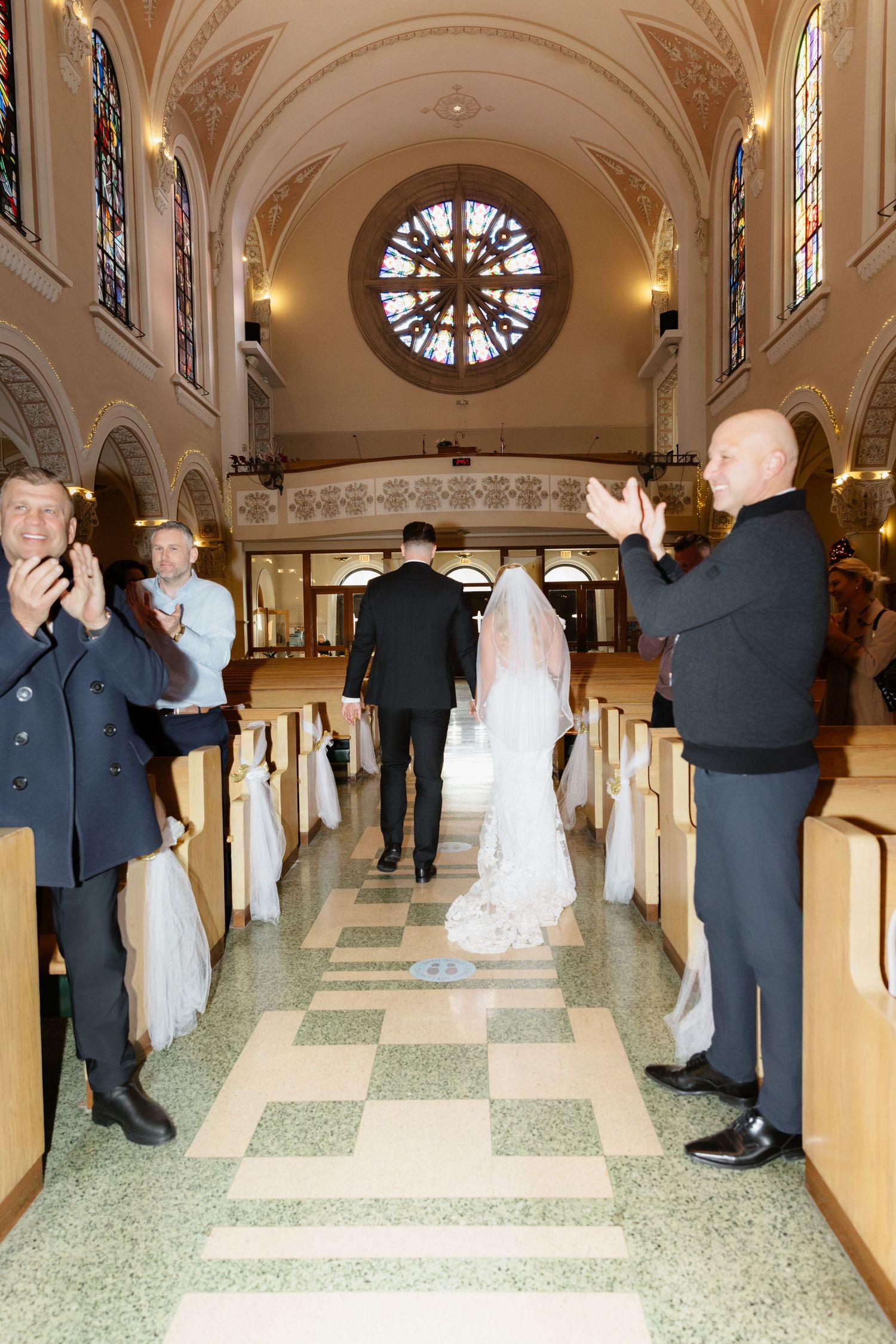 Guests clap as newlyweds walk down the aisle of a church with stained glass rose window.