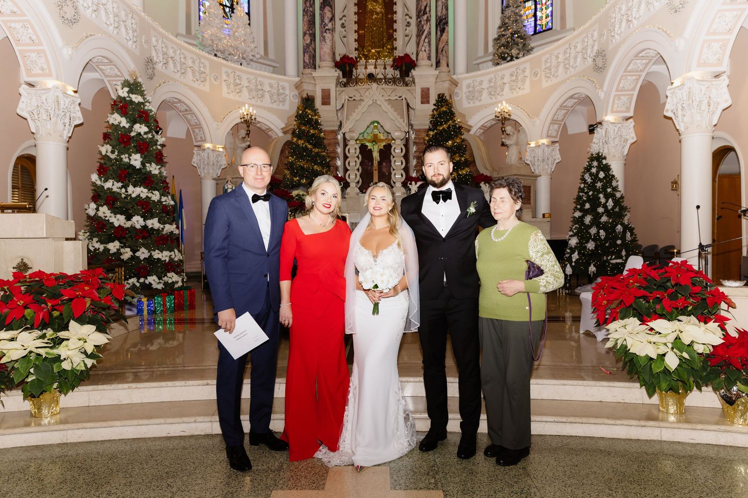 Bride and groom with parents posing in a Christmas-decorated Catholic church surrounded by poinsettias and holiday trees.