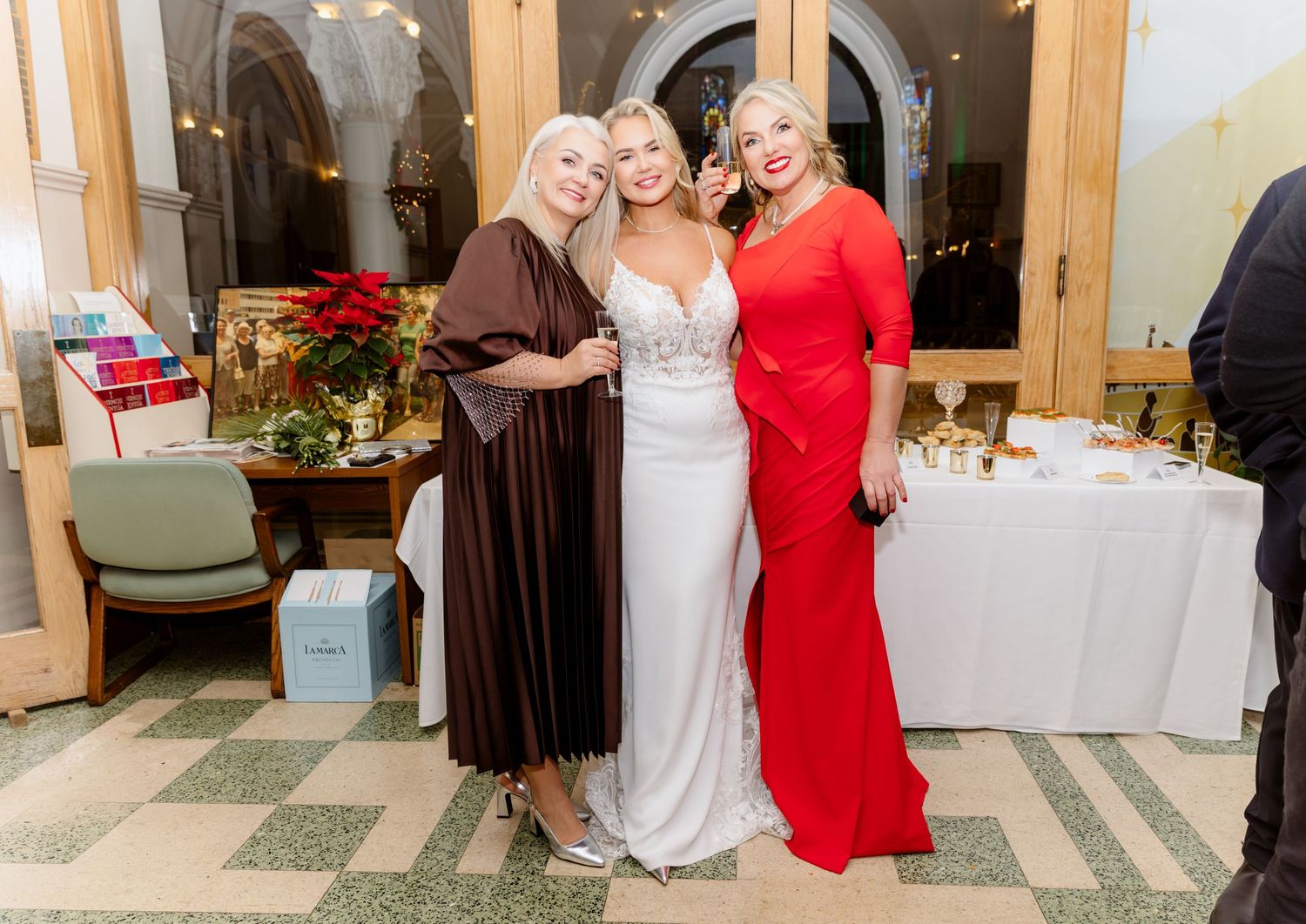 Bride in white gown poses with two women in formal attire at a holiday wedding reception.