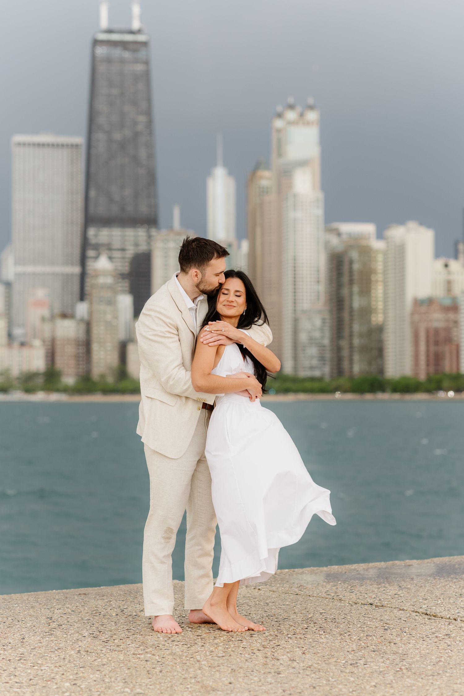 Couple sharing romantic kiss on Chicago waterfront pier, woman in white dress with John Hancock Tower in background.