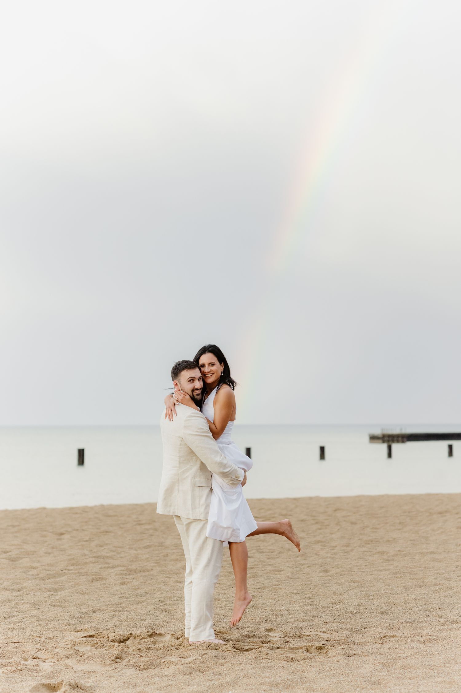 A couple hugging on a sandy beach with a rainbow and ocean pier visible in the background.