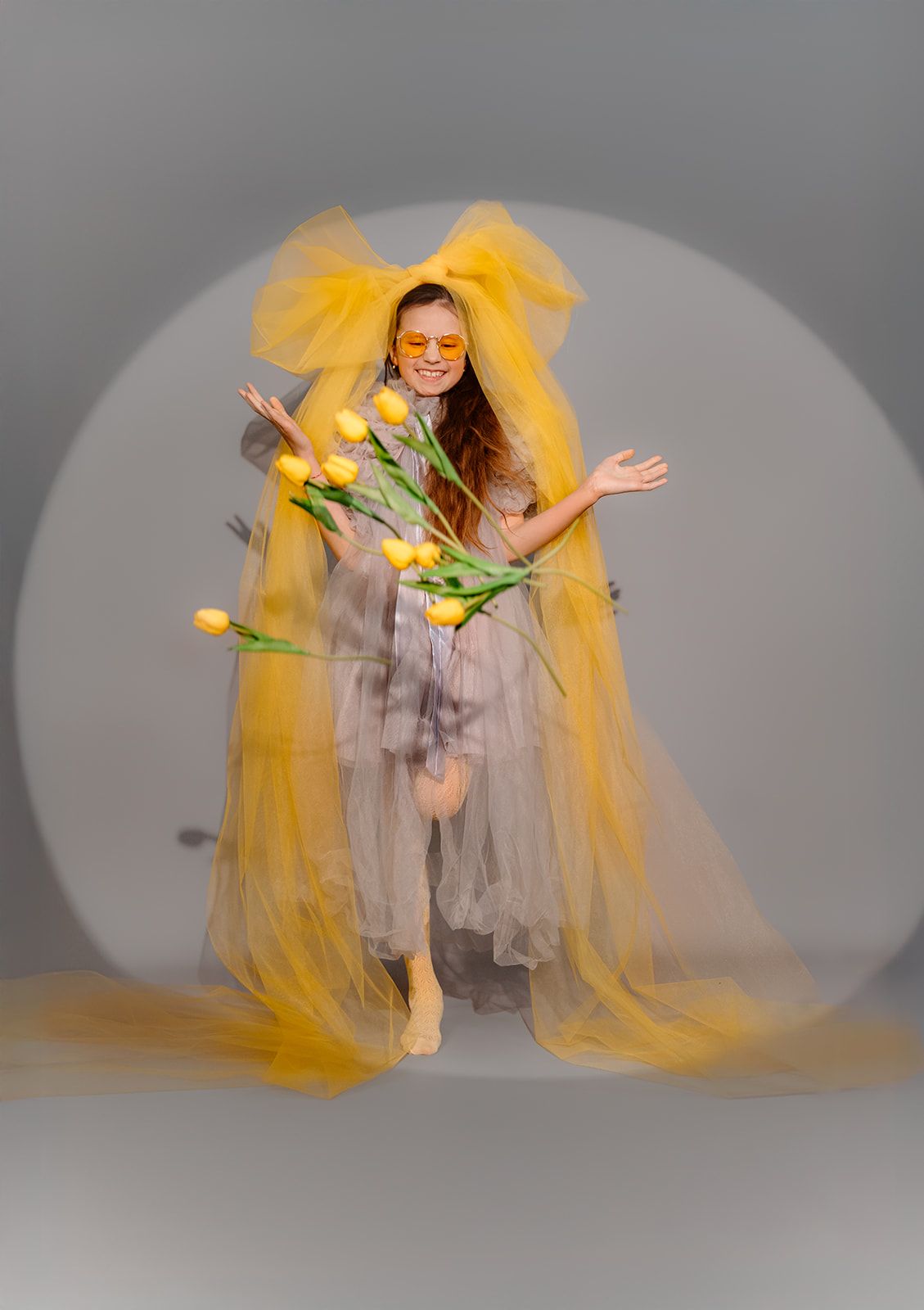 Woman in yellow tulle gown holding yellow tulips, jumping joyfully against a gray studio background.