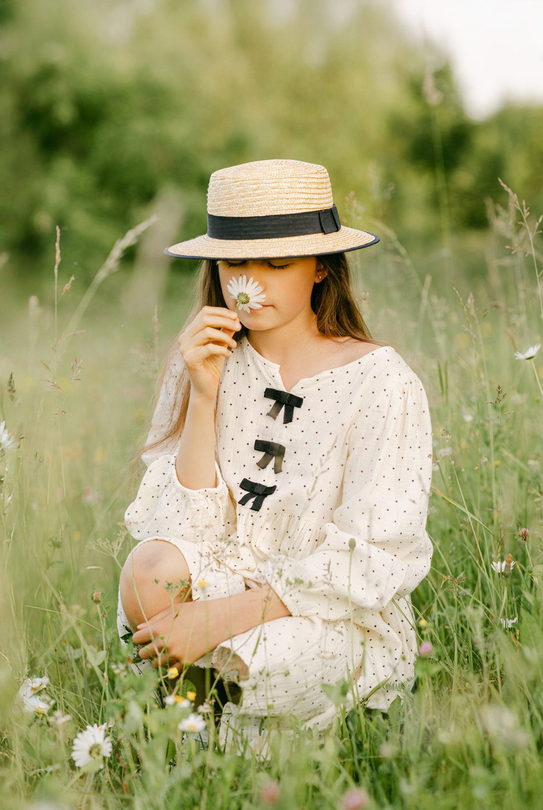 Young girl in white polka dot dress and straw hat smells daisy flower while sitting in meadow.