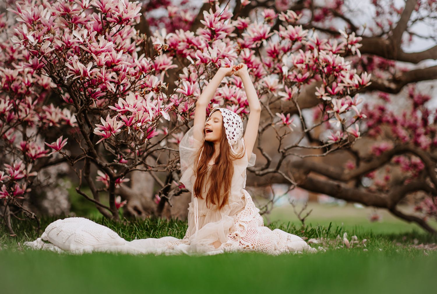 Woman in white dress sits gracefully under blooming pink magnolia trees on green grass, arms raised elegantly.